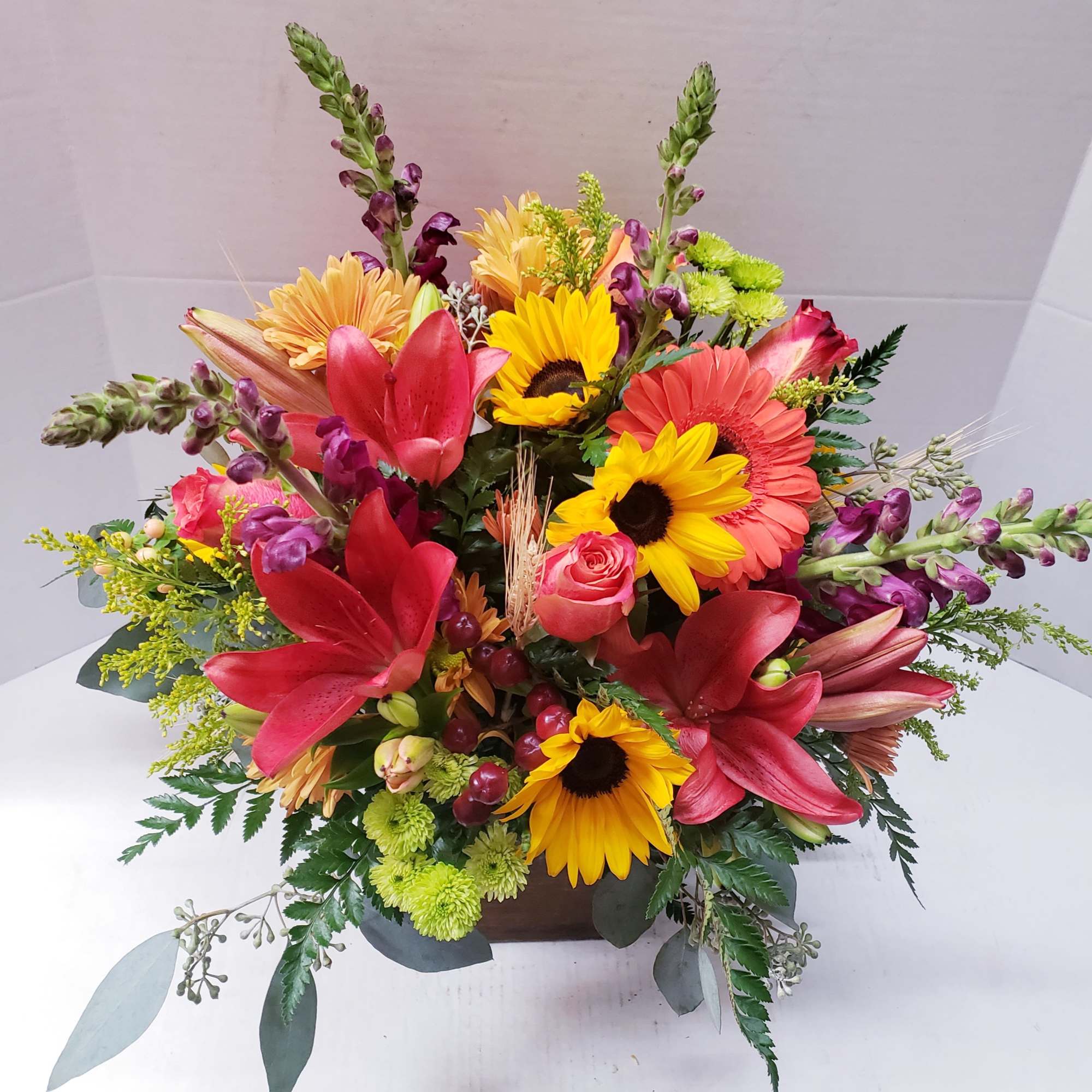 Bright arrangement of red lilies, yellow sunflowers, roses, and mixed blooms in a wooden box.