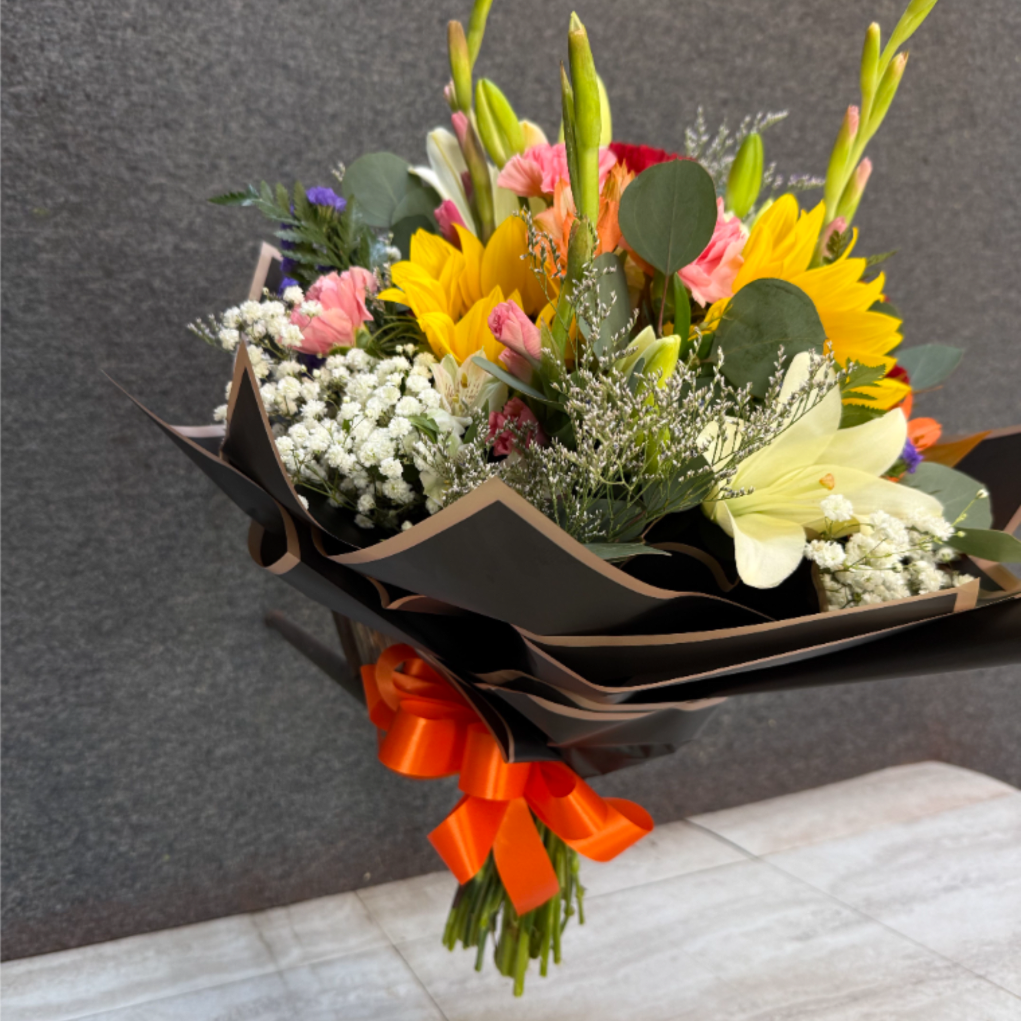 Hand-tied bouquet with yellow sunflowers, white lilies, and pink flowers wrapped in black paper with an orange bow.