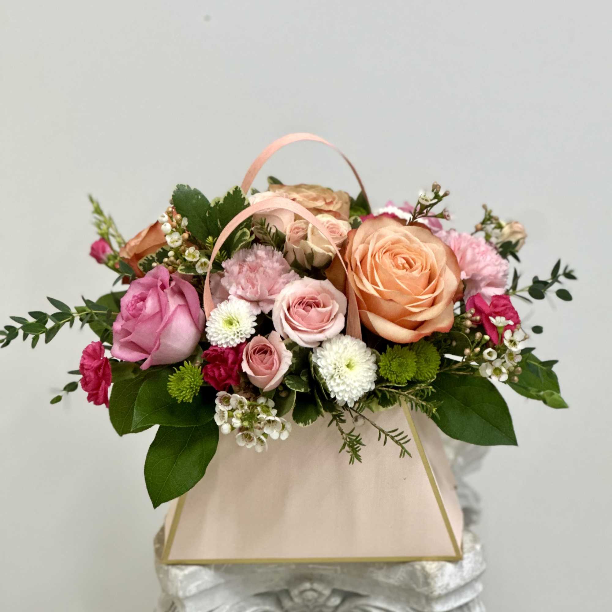 Pastel pink and peach flower arrangement with roses and mums in a blush gift bag-style container.