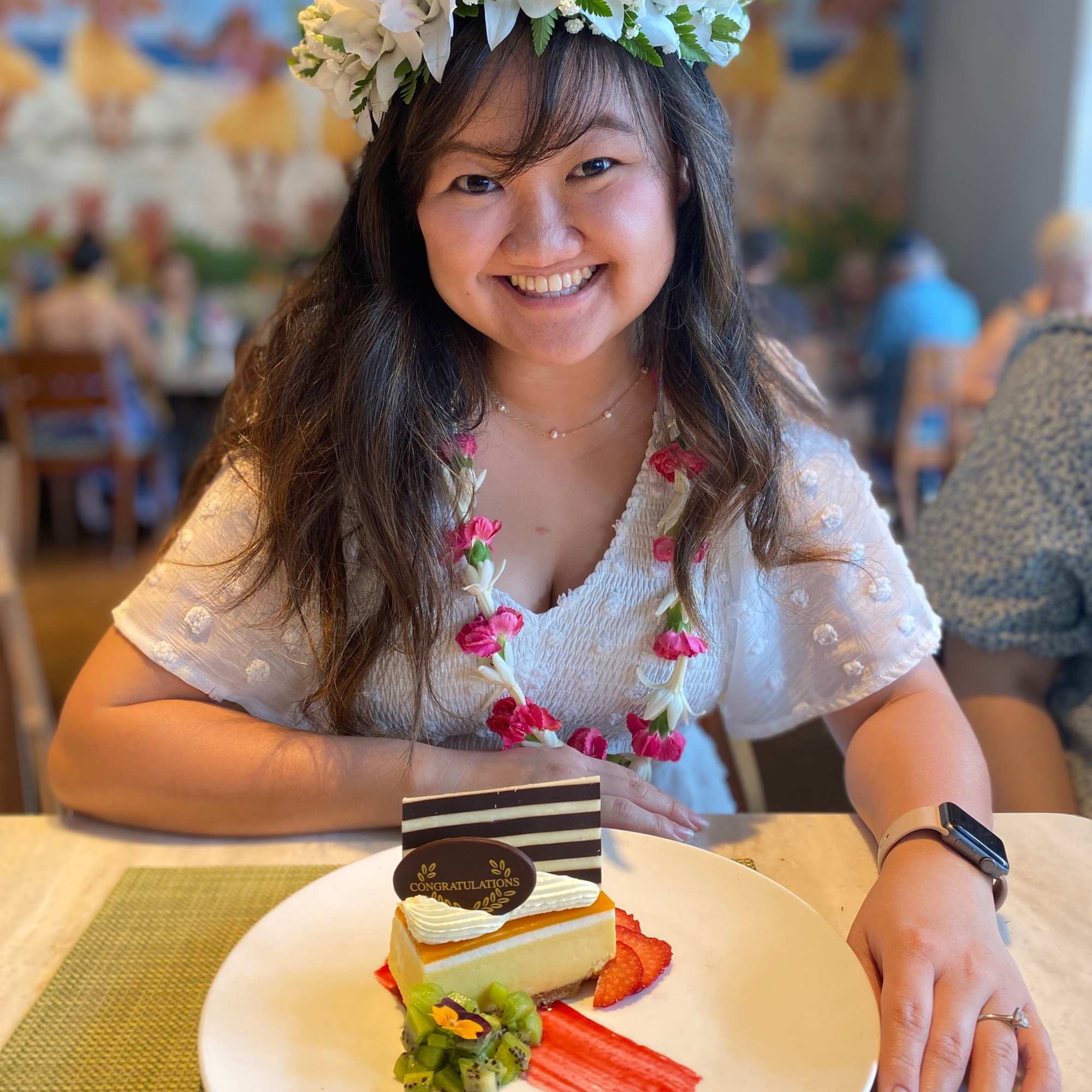 Woman wearing a white flower crown and floral lei sitting with a congratulatory dessert plate