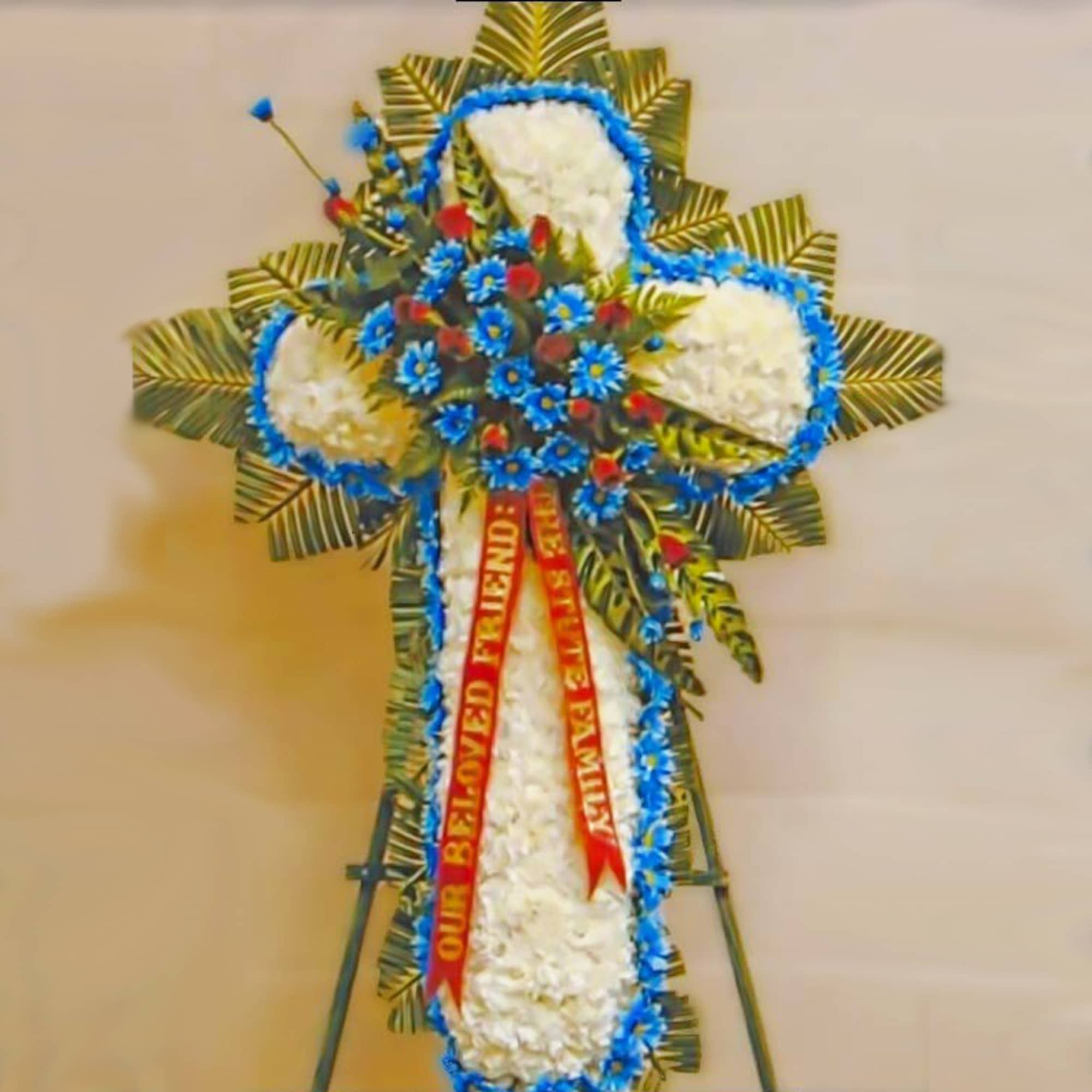 Standing white cross floral spray with blue accents, palm fronds, and red roses on an easel with memorial ribbon