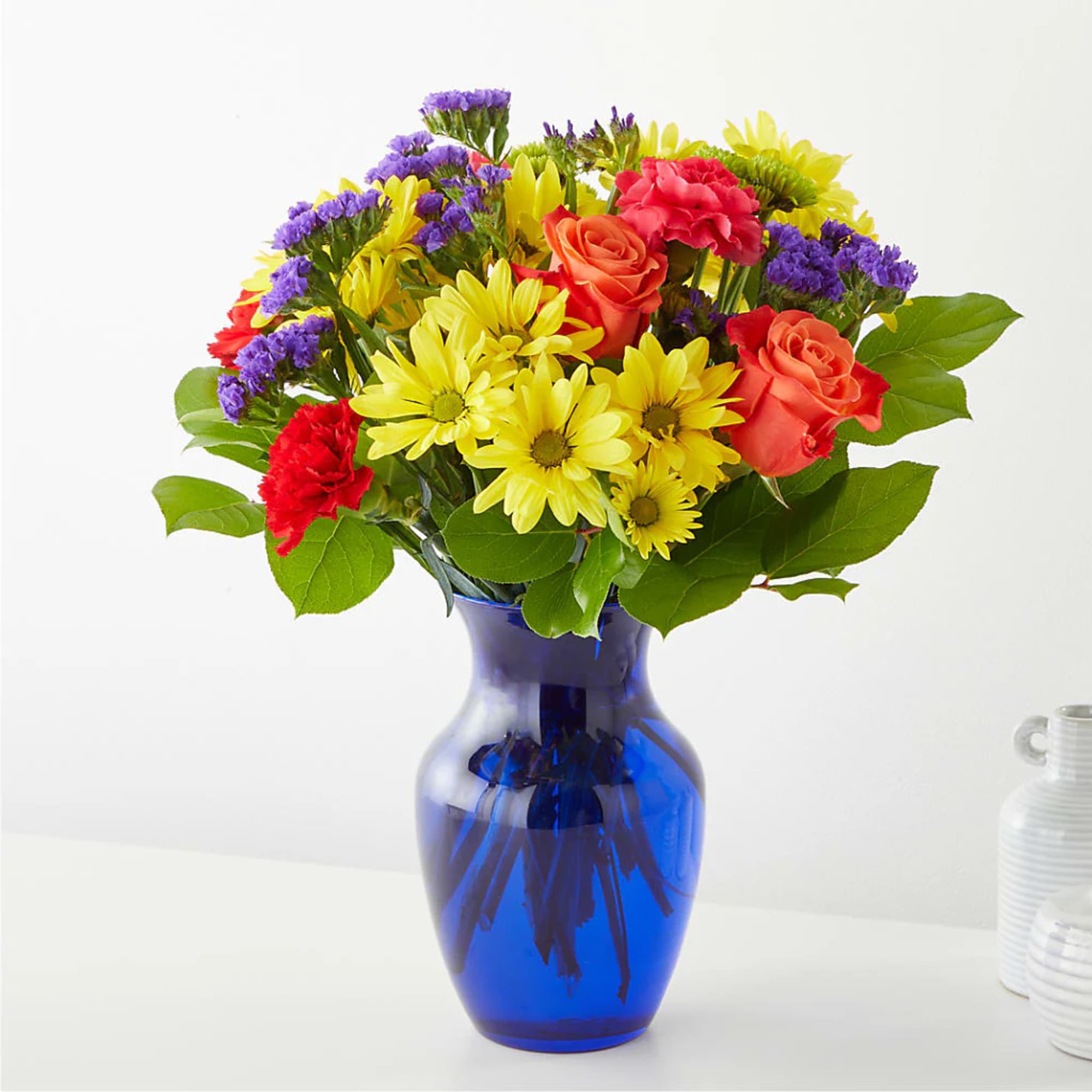 Mixed bouquet of yellow daisies, orange roses, red carnations, and purple filler in a blue vase