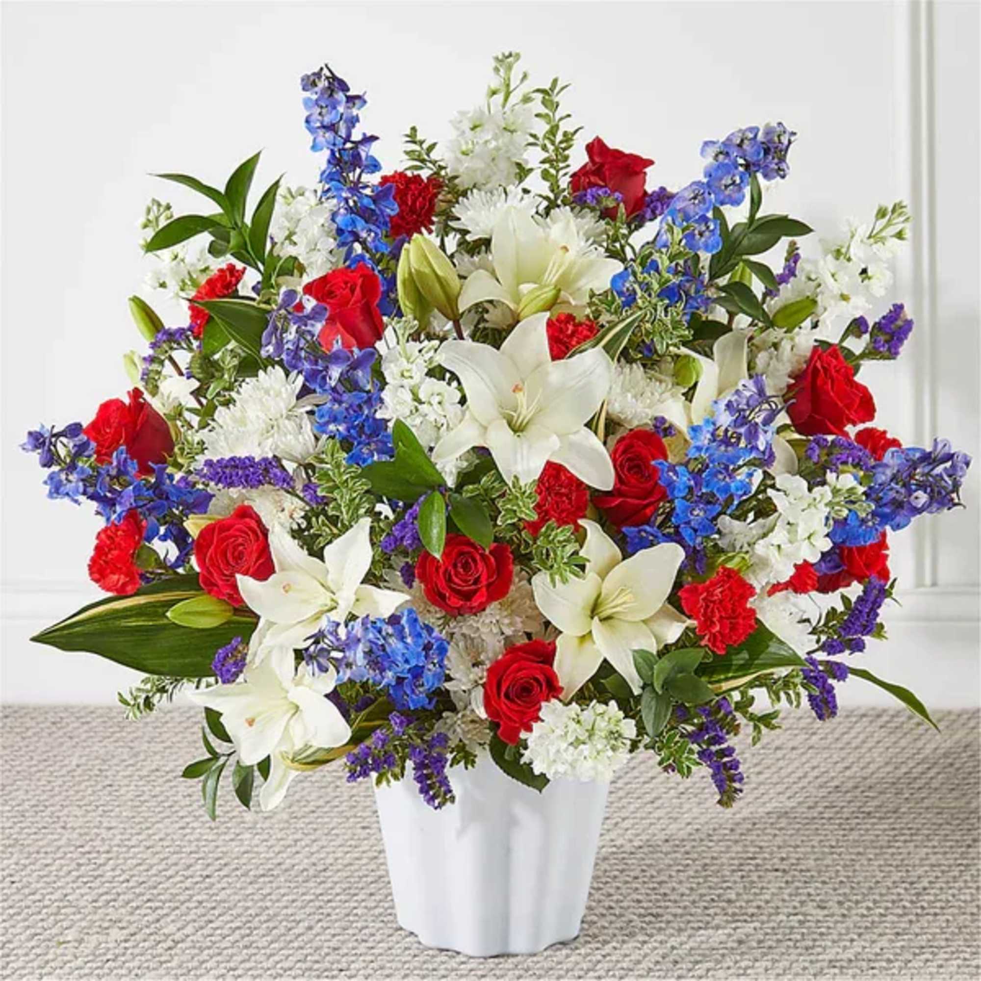 Red roses, white lilies, and blue flowers arranged in a white fluted vase.