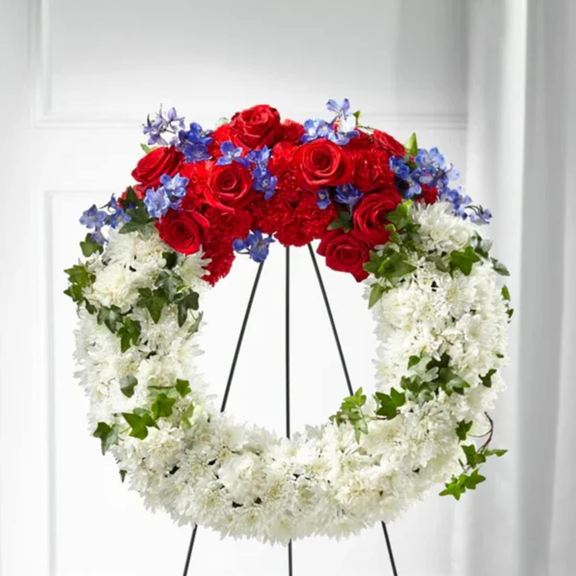 Heart-shaped floral wreath with red roses, white flowers, and blue accents on a stand