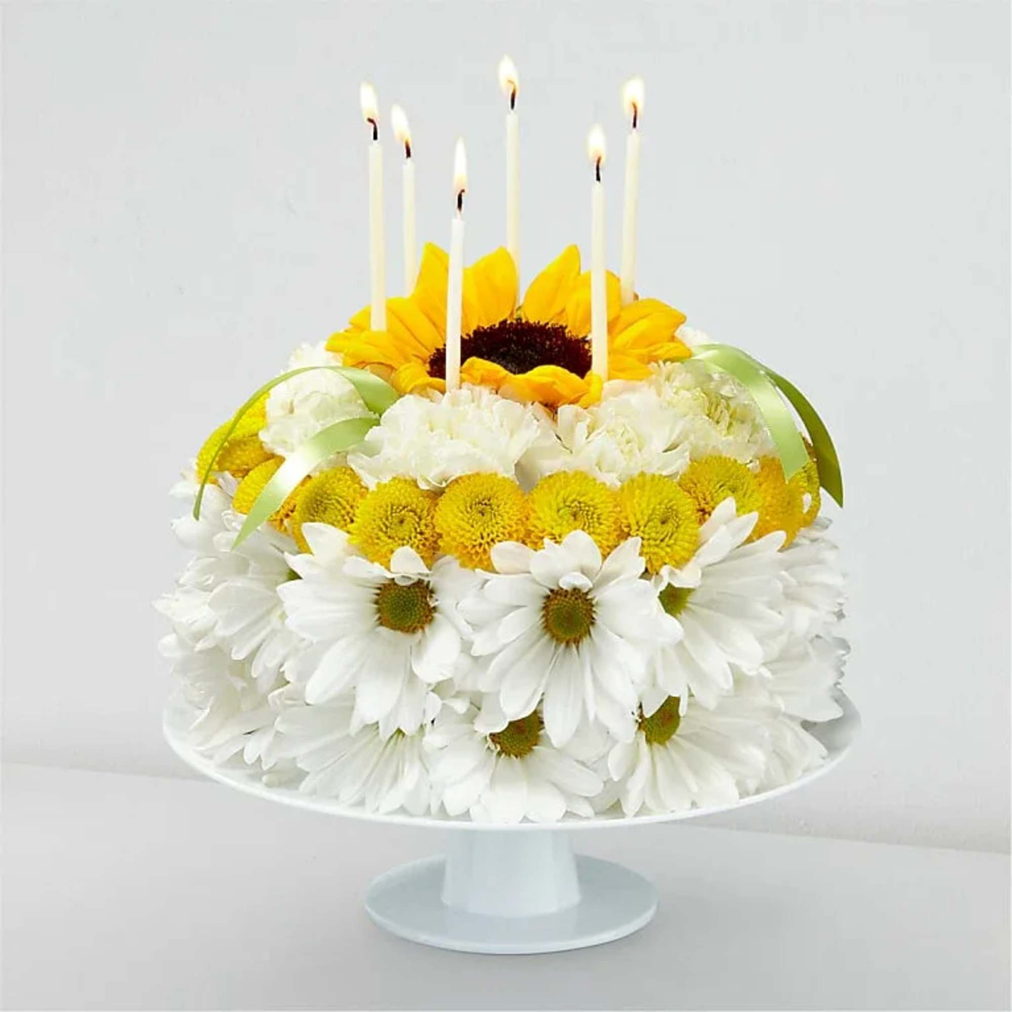 Flower cake with white daisies, yellow blooms, and lit candles on a pedestal stand