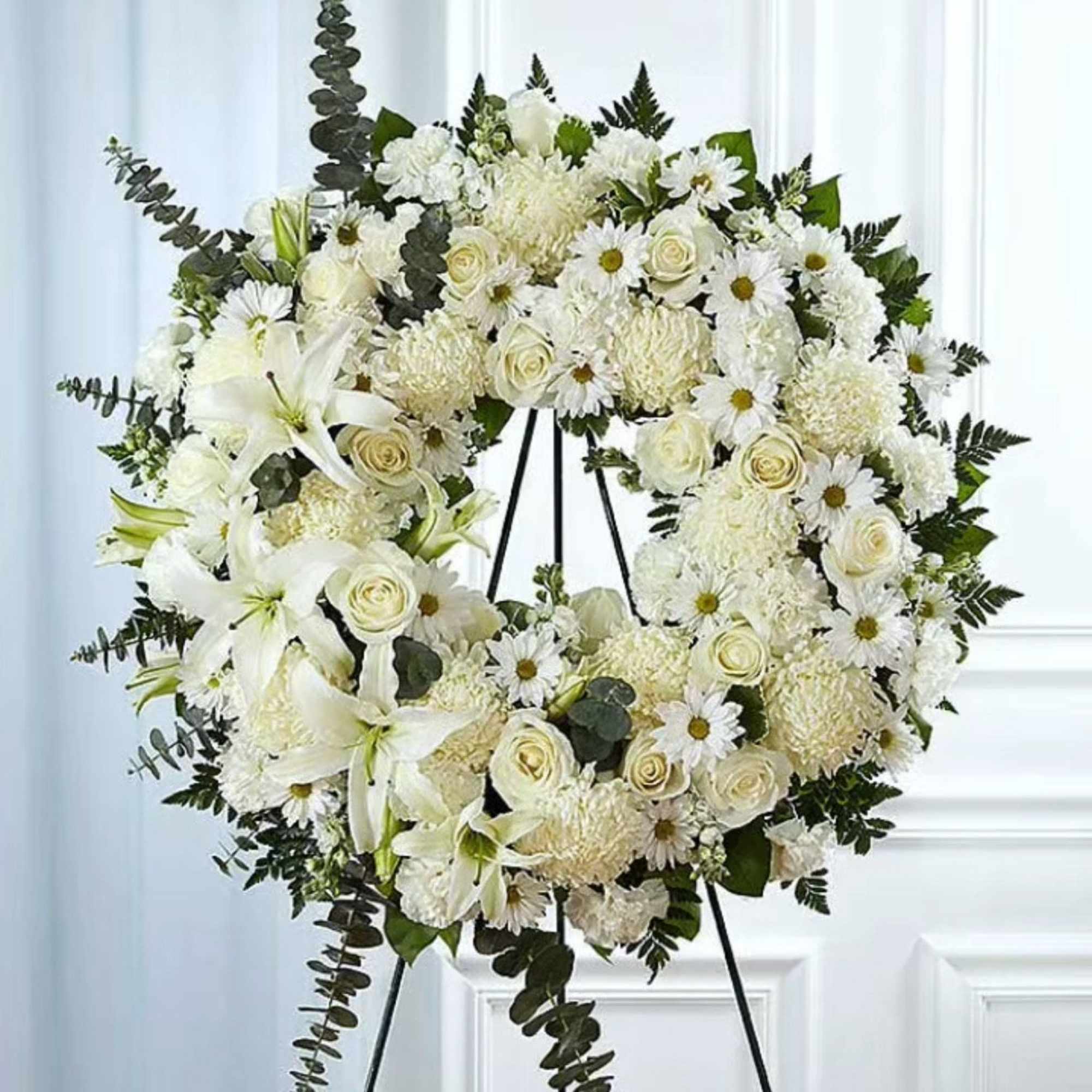 White floral wreath on a black easel with lilies, roses, and daisies