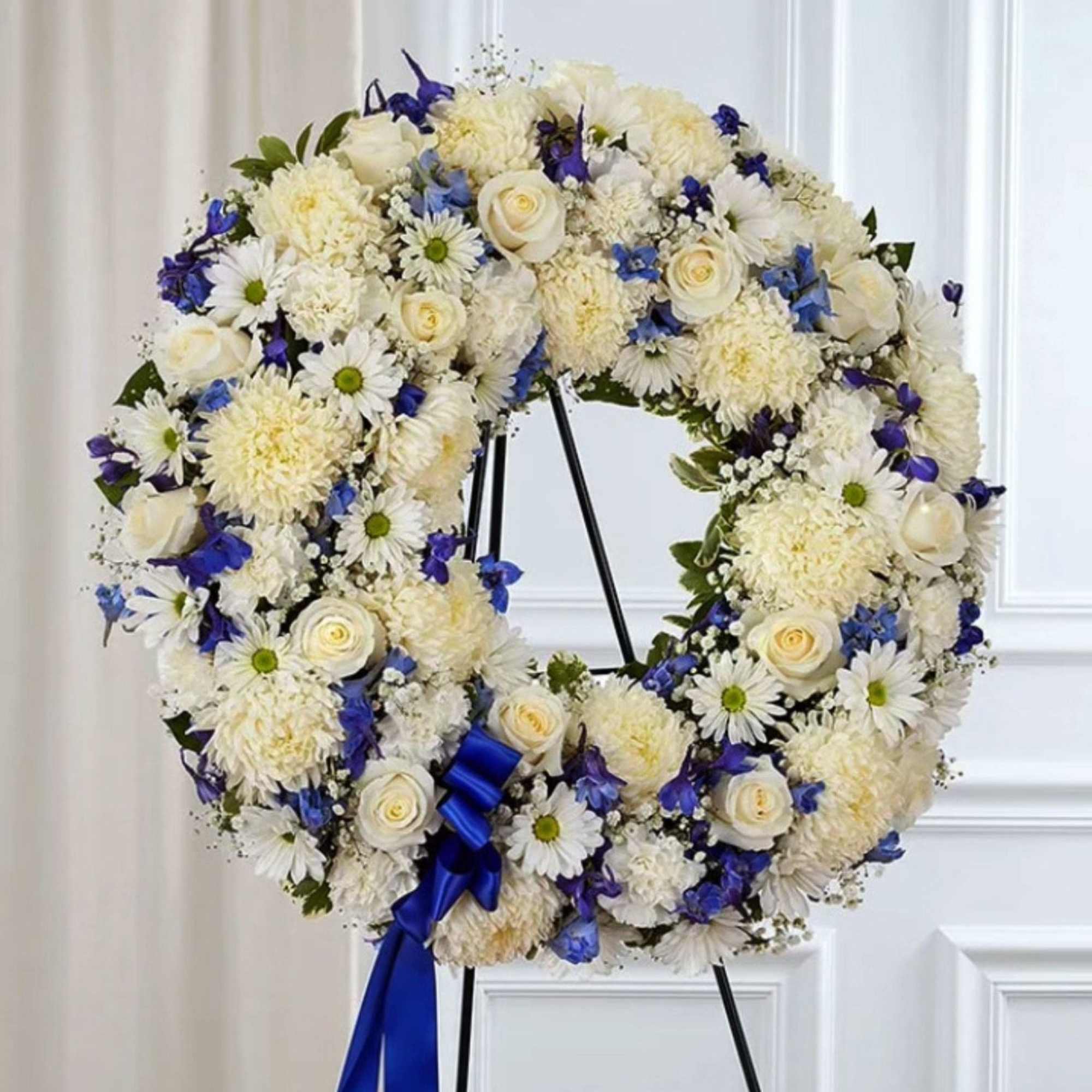 White floral wreath with blue accents on a black stand and ribbon