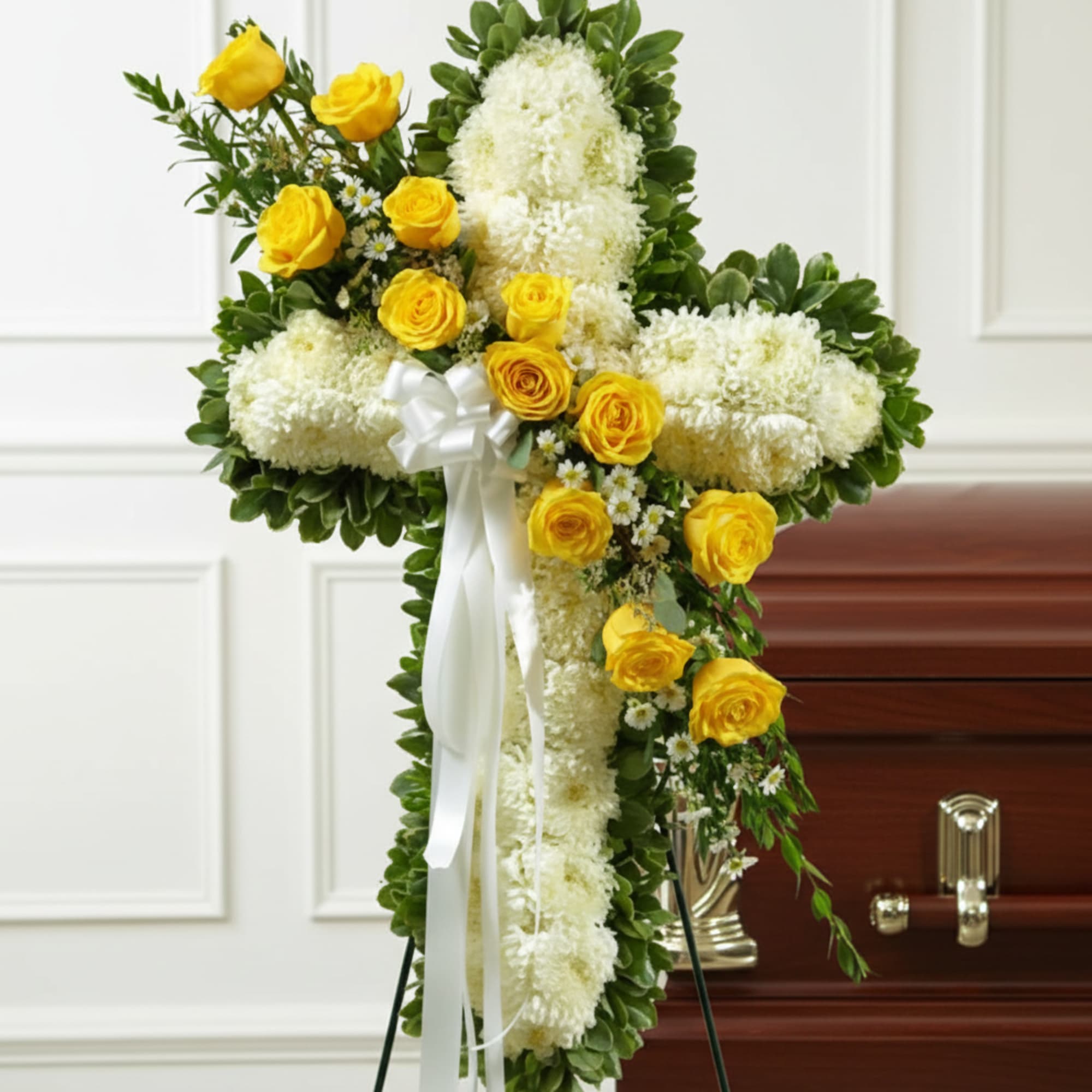White floral cross with yellow roses and a white ribbon on a stand