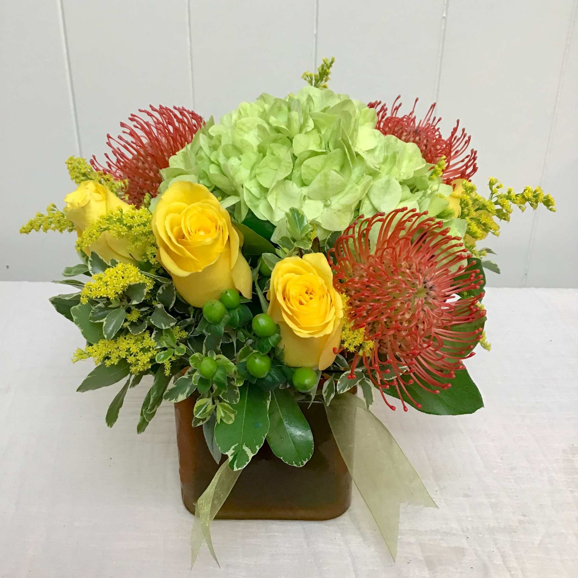 Compact arrangement of yellow roses, green hydrangea, and orange pincushion protea in a cube vase.