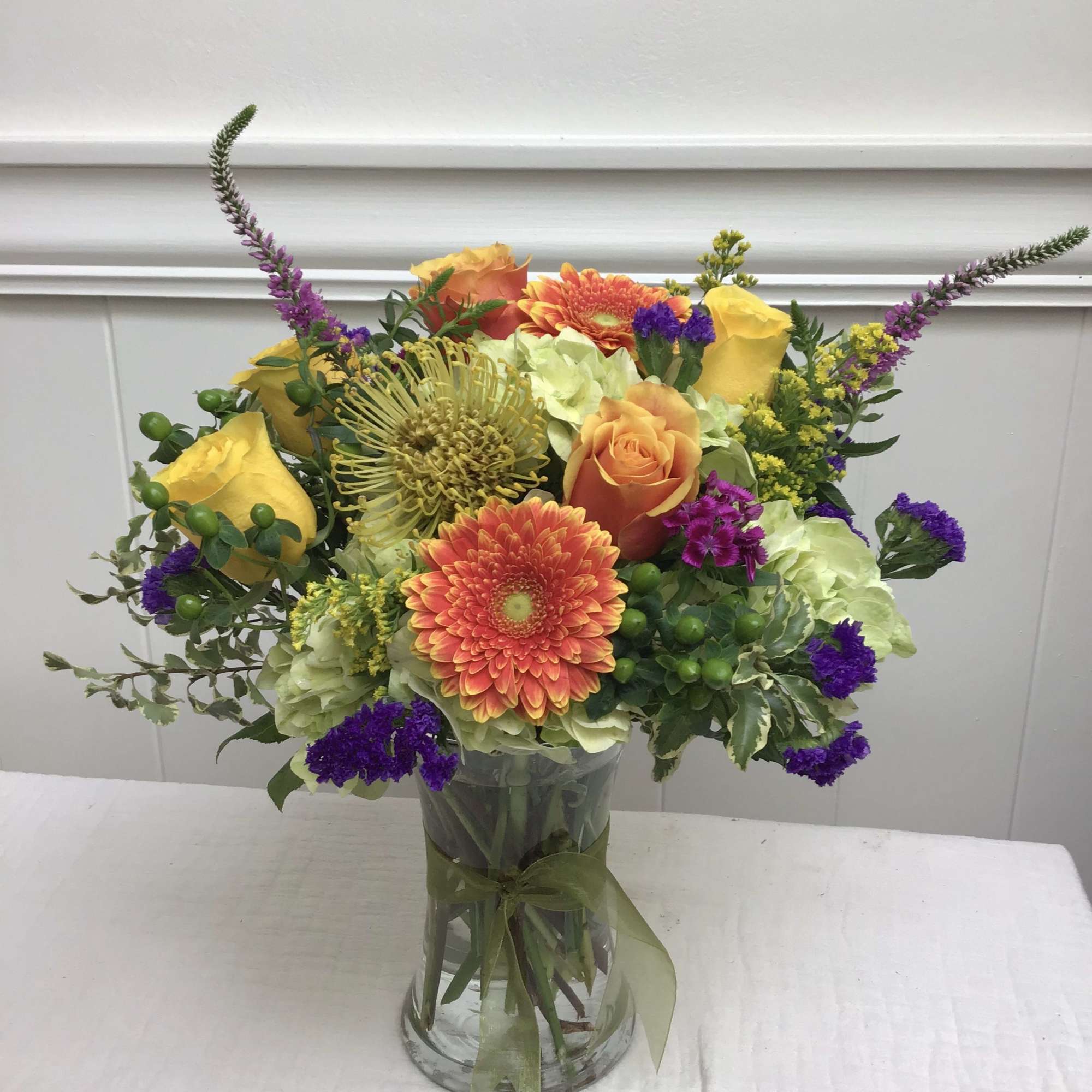 Bright mixed bouquet of yellow roses, orange gerbera daisies, and purple flowers in a clear glass vase.
