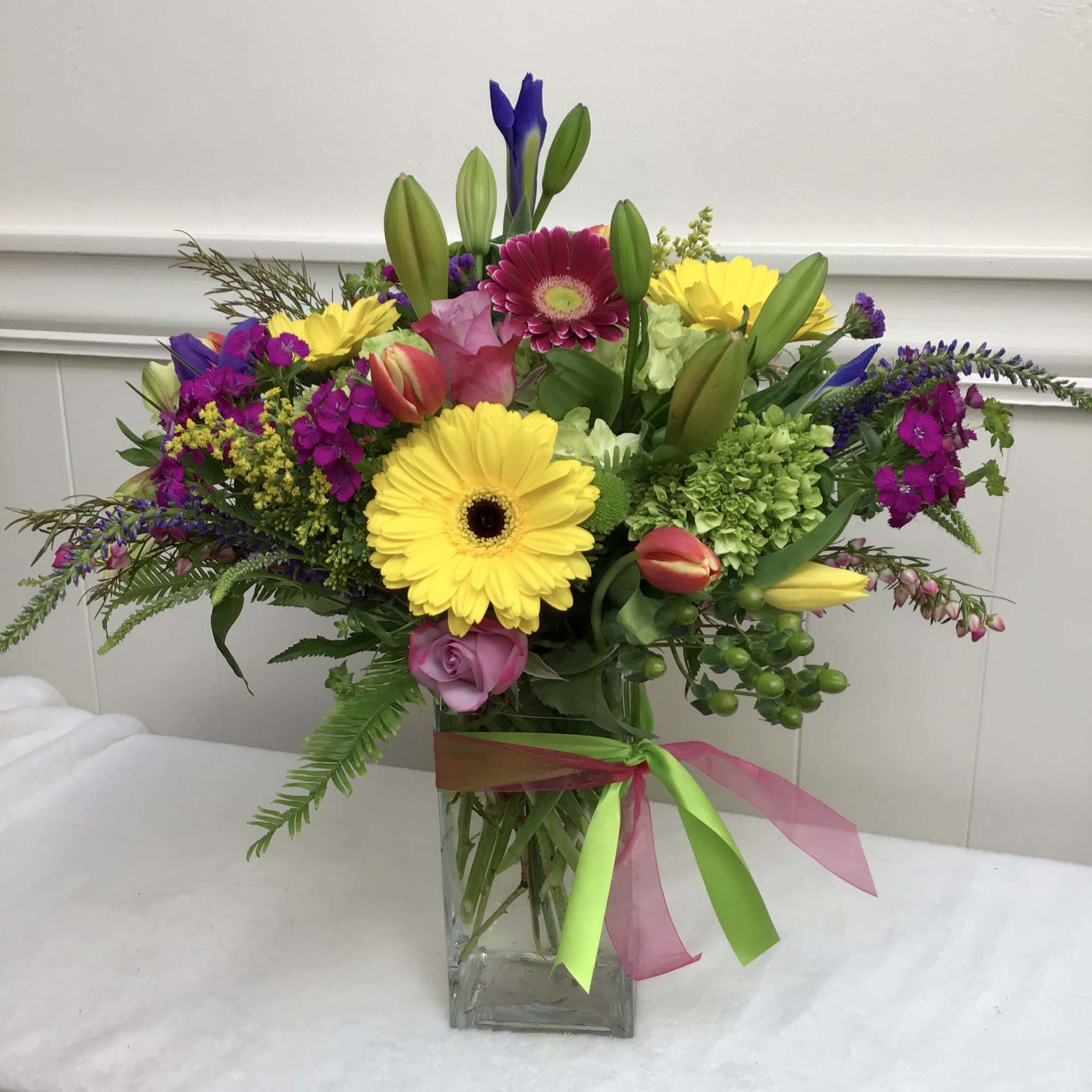 Bright mixed bouquet with yellow gerbera daisies, roses, tulips and lilies in a clear rectangular vase with ribbon