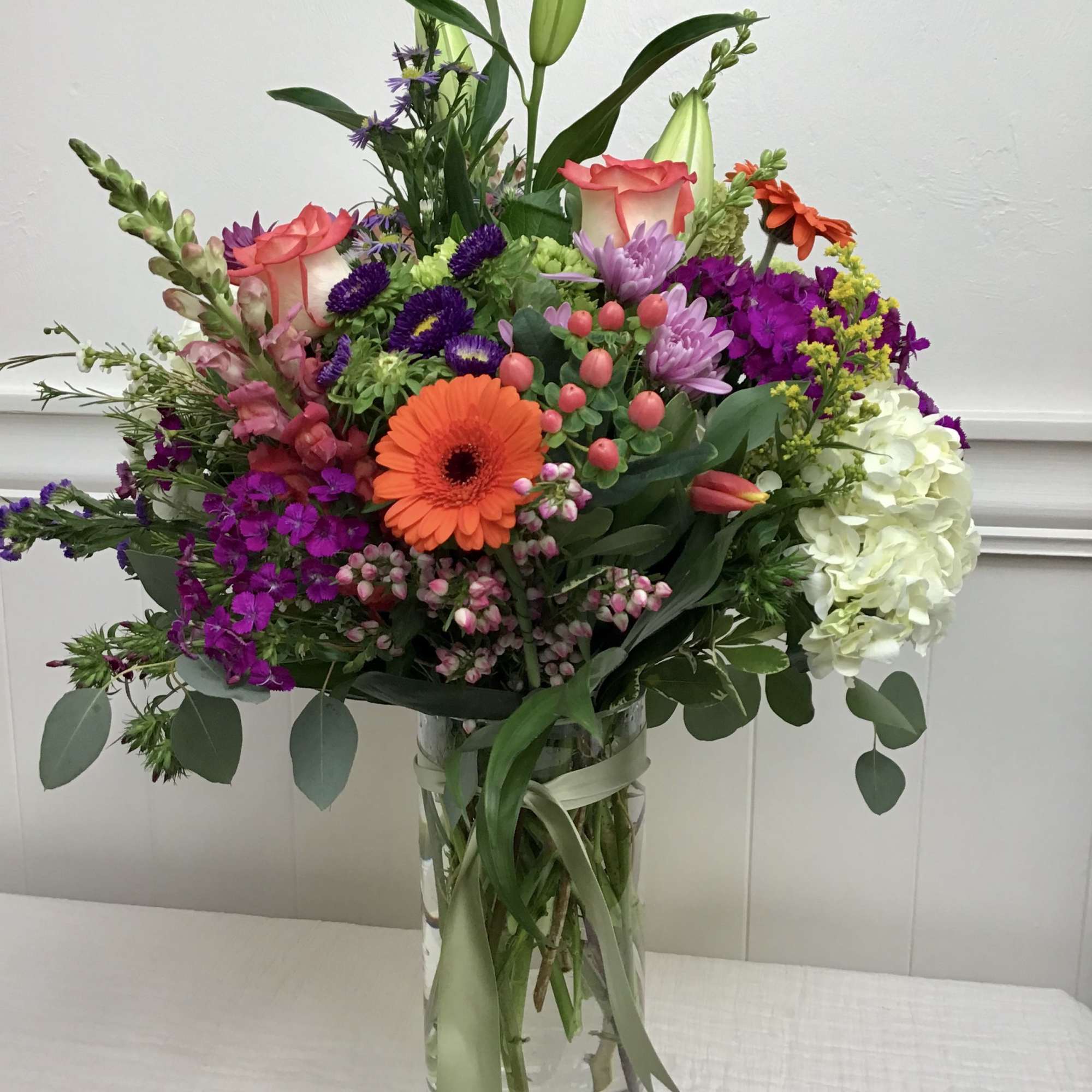 Tall mixed bouquet with orange gerbera, pink roses, lilies, and white hydrangea in a clear glass vase