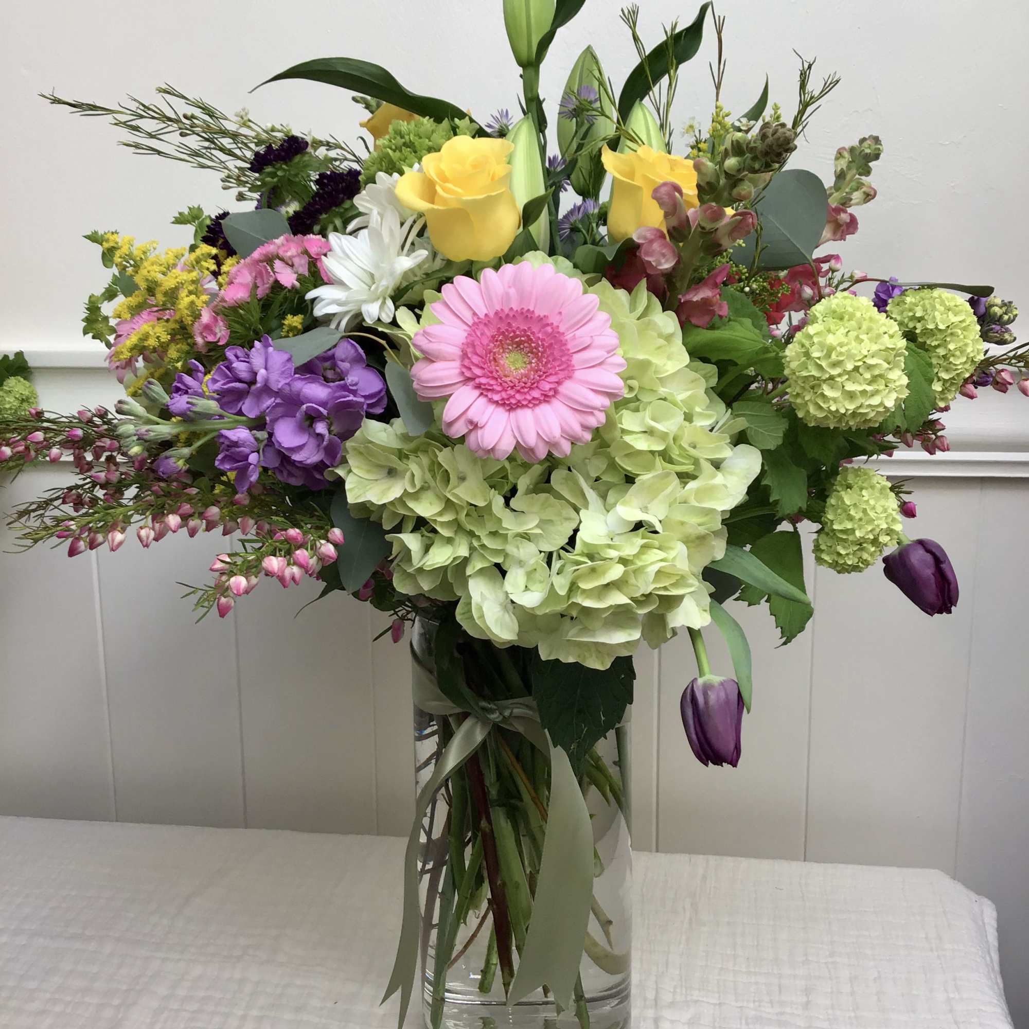 Tall mixed arrangement with pink gerbera, yellow roses, green hydrangeas and purple tulips in a glass vase