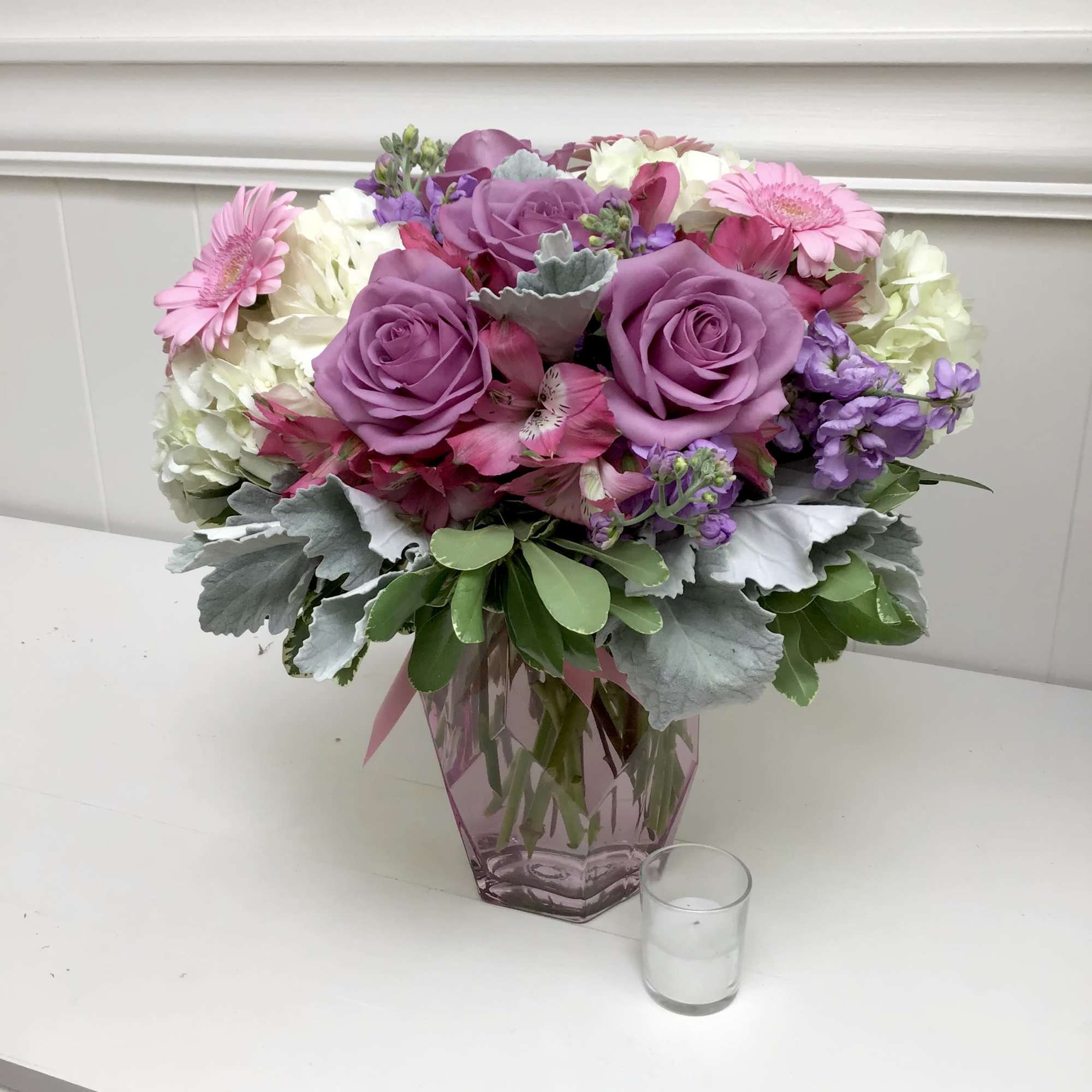 Pink and purple roses, gerberas, hydrangeas and mixed blooms in a pink glass vase on a white table.