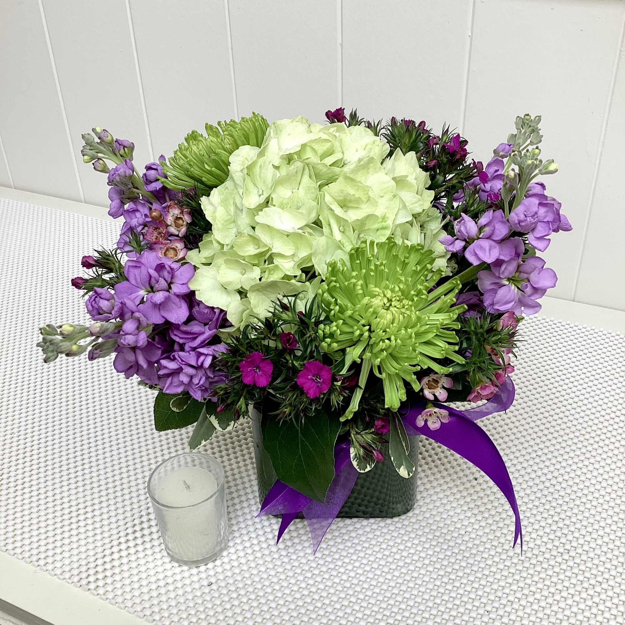 Compact arrangement of green hydrangea and mums with purple blooms in a cube vase beside a glass votive candle