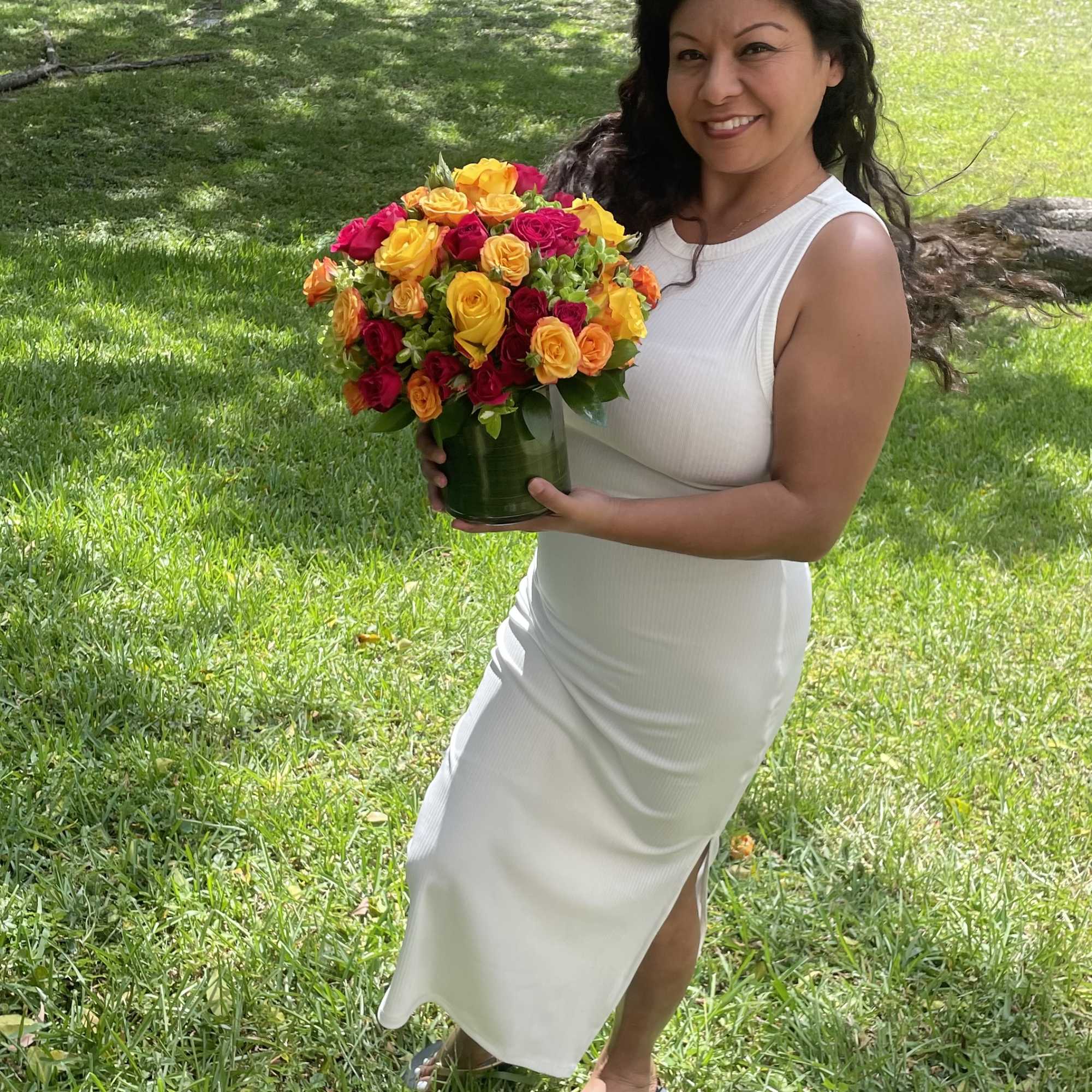 Woman holding a bouquet of yellow, orange, and pink roses in a glass vase