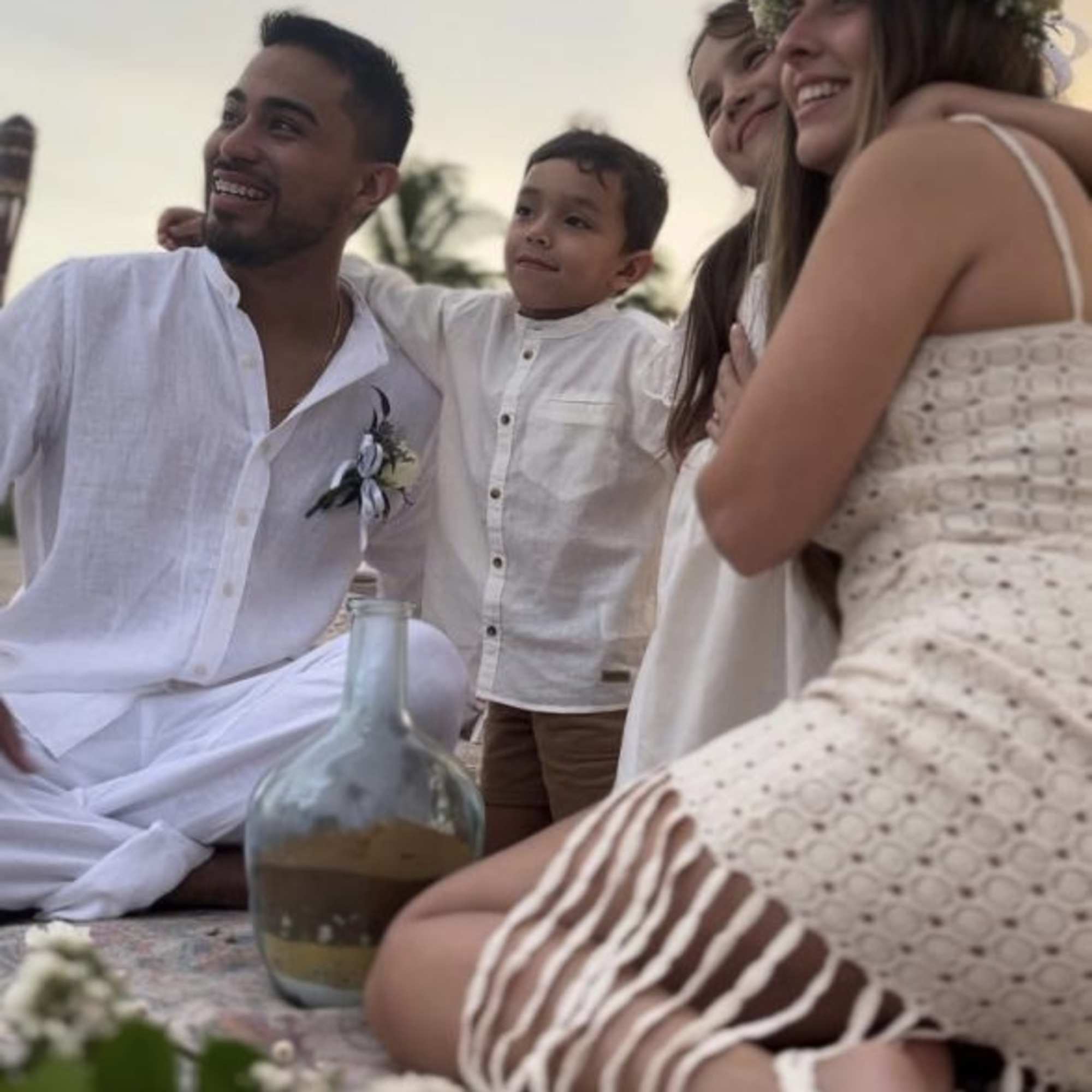 People in white clothing sit outdoors with a floral crown and small flowers in the foreground.