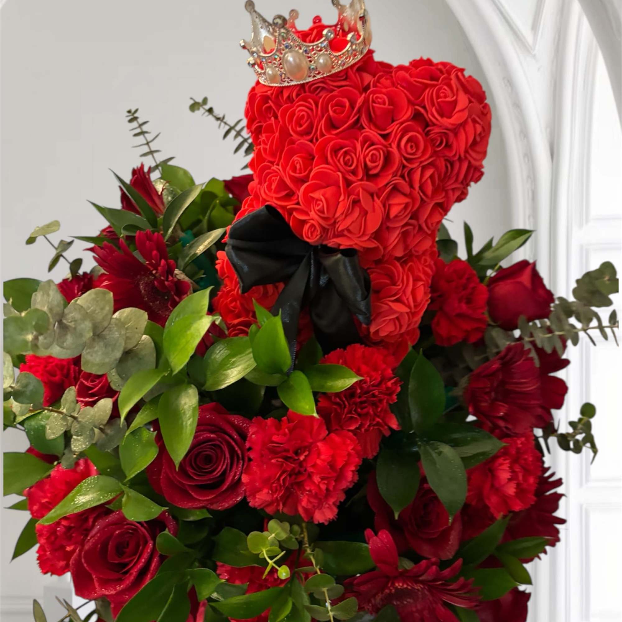Red rose teddy bear with a crown and black bow, surrounded by red flowers