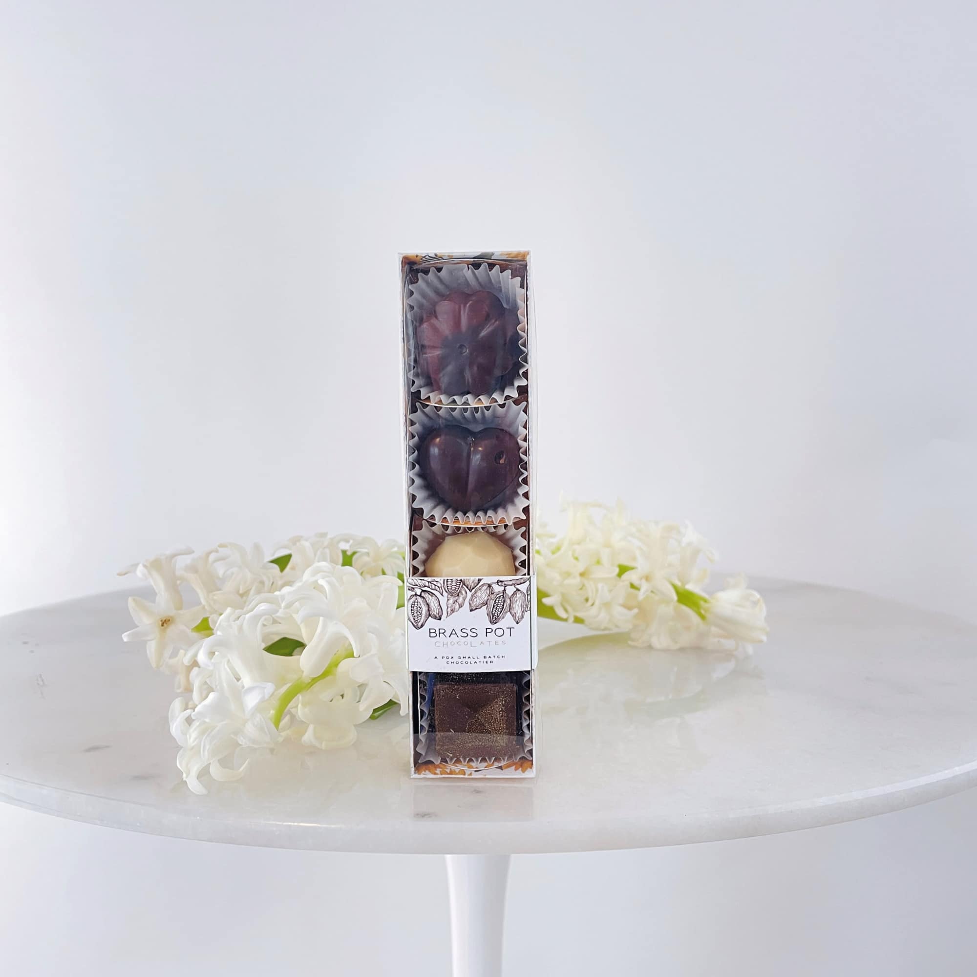 Box of chocolates on a white table with white flowers behind it