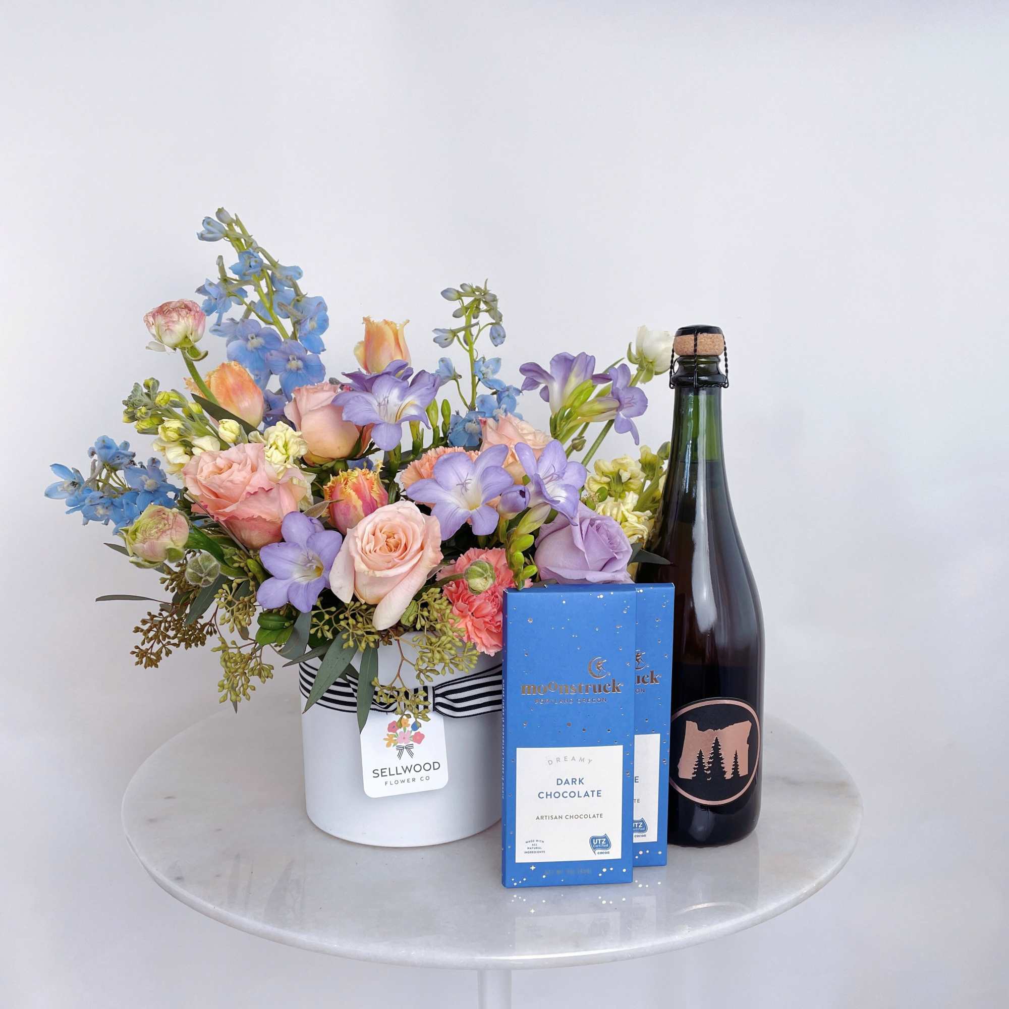 Mixed pastel flower arrangement in a white hatbox with chocolate and a bottle of wine