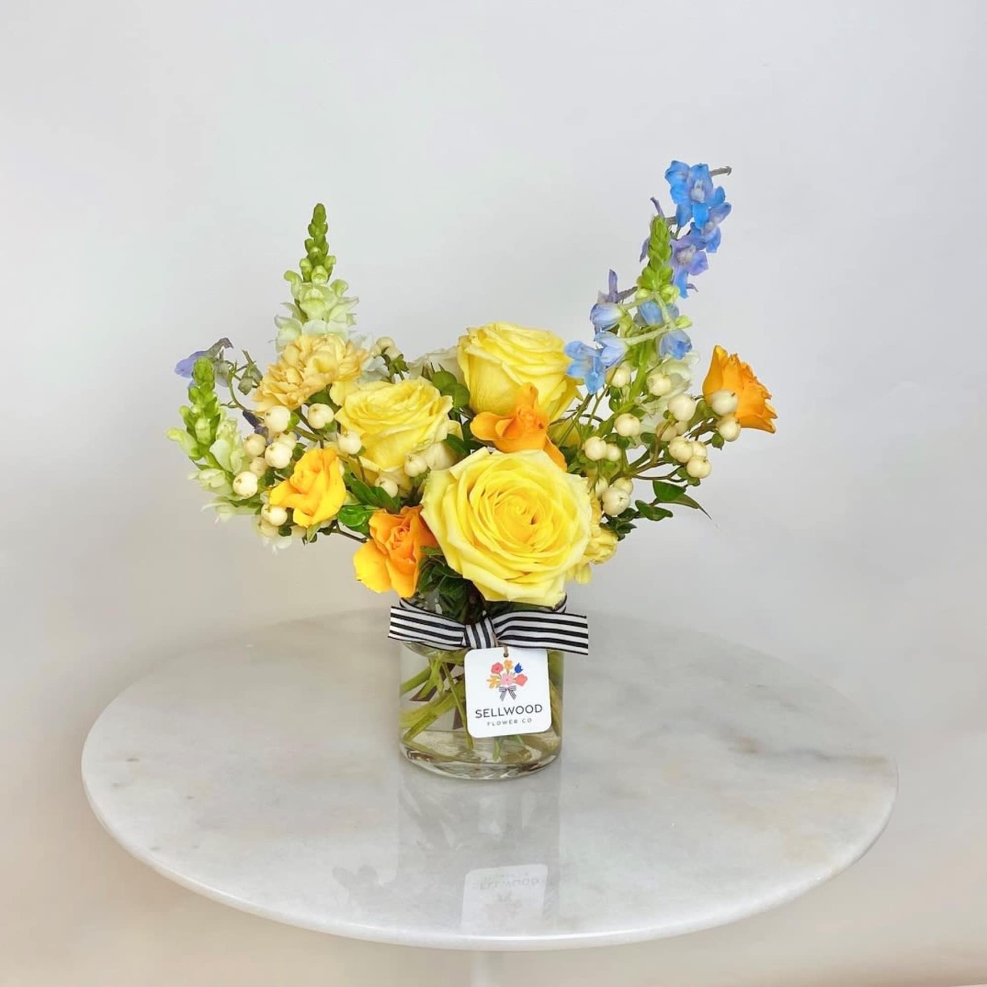 Yellow and orange rose arrangement with blue delphinium in a glass jar with striped ribbon