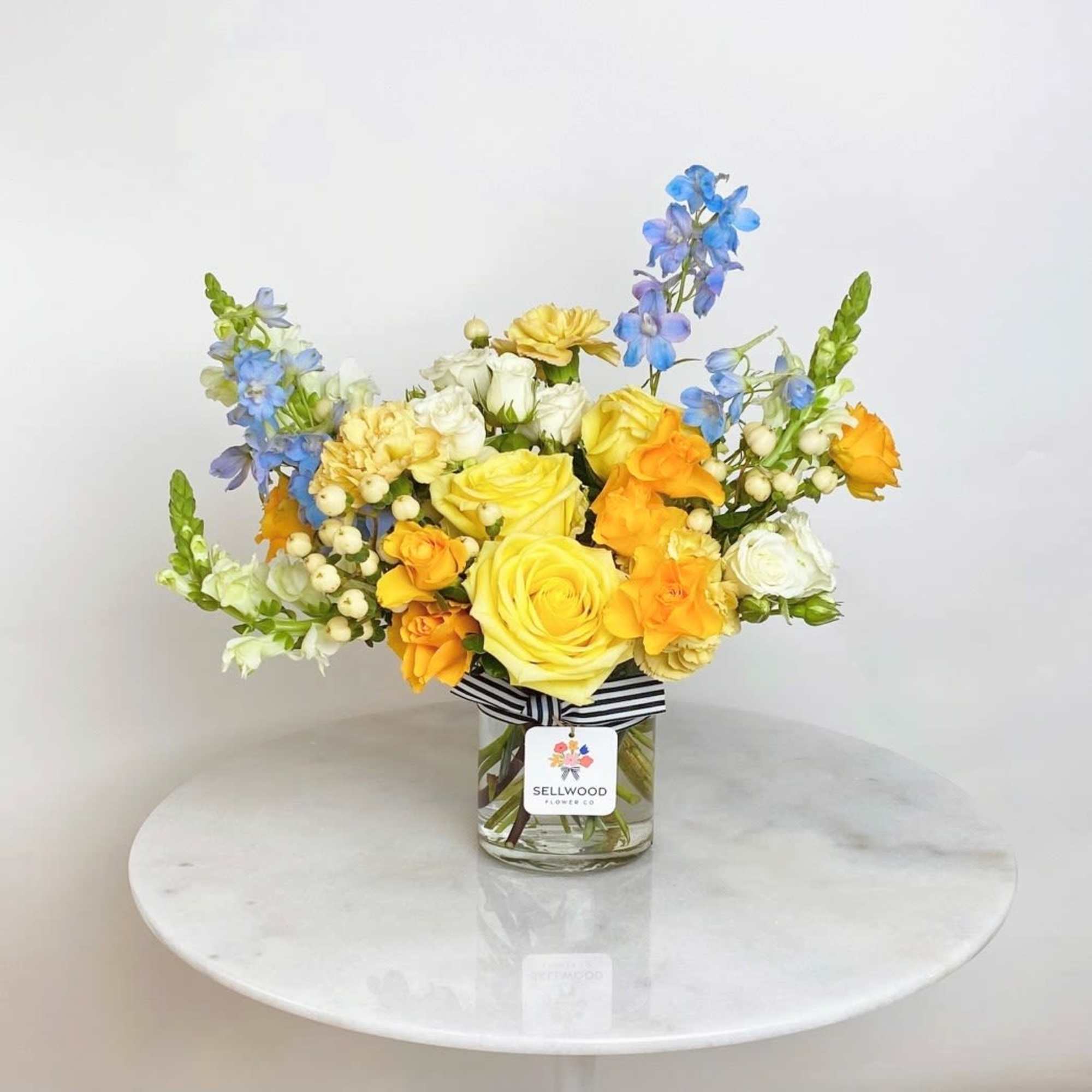 Yellow and orange roses with blue delphinium in a clear glass vase on a round white table