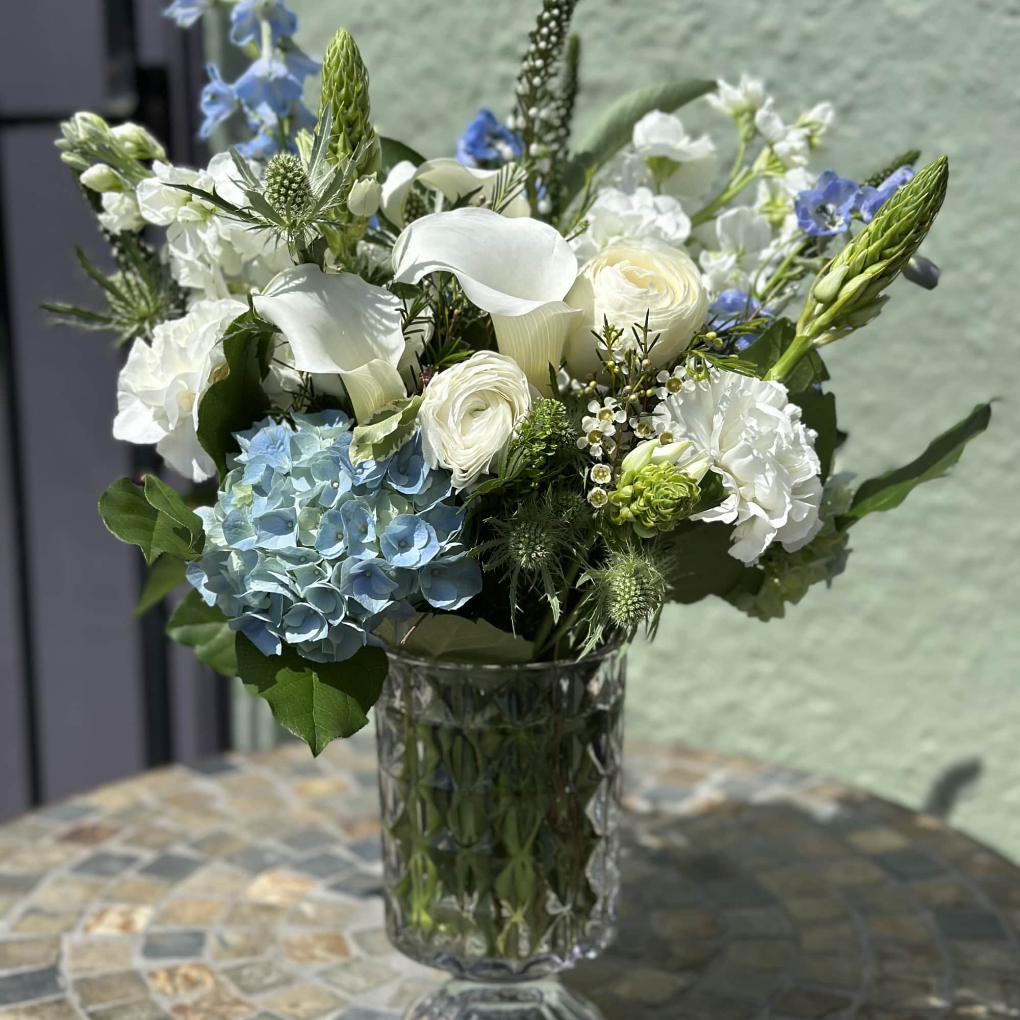 White and blue flower arrangement with calla lilies and hydrangeas in a tall patterned glass vase