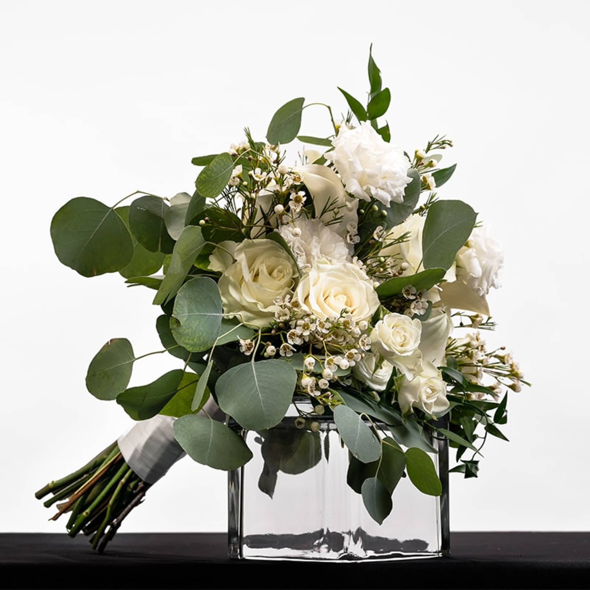 Hand-tied bouquet of white roses and blooms with lush round foliage in a clear glass cube vase