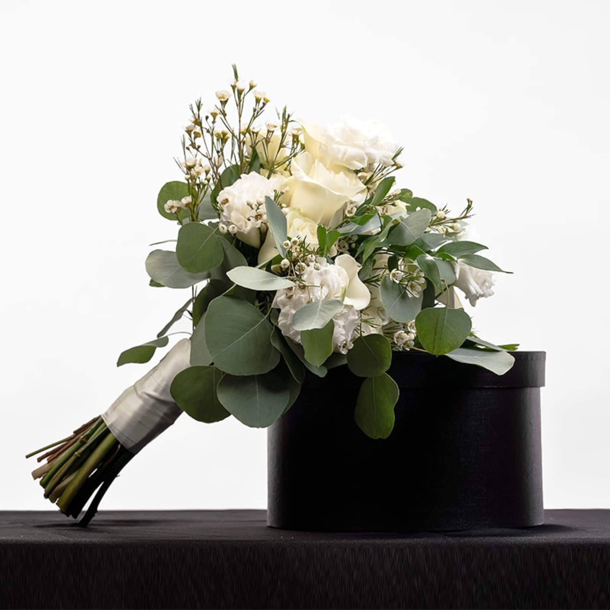 Hand-tied bouquet of white roses and small white blooms with eucalyptus, resting on a black hatbox.