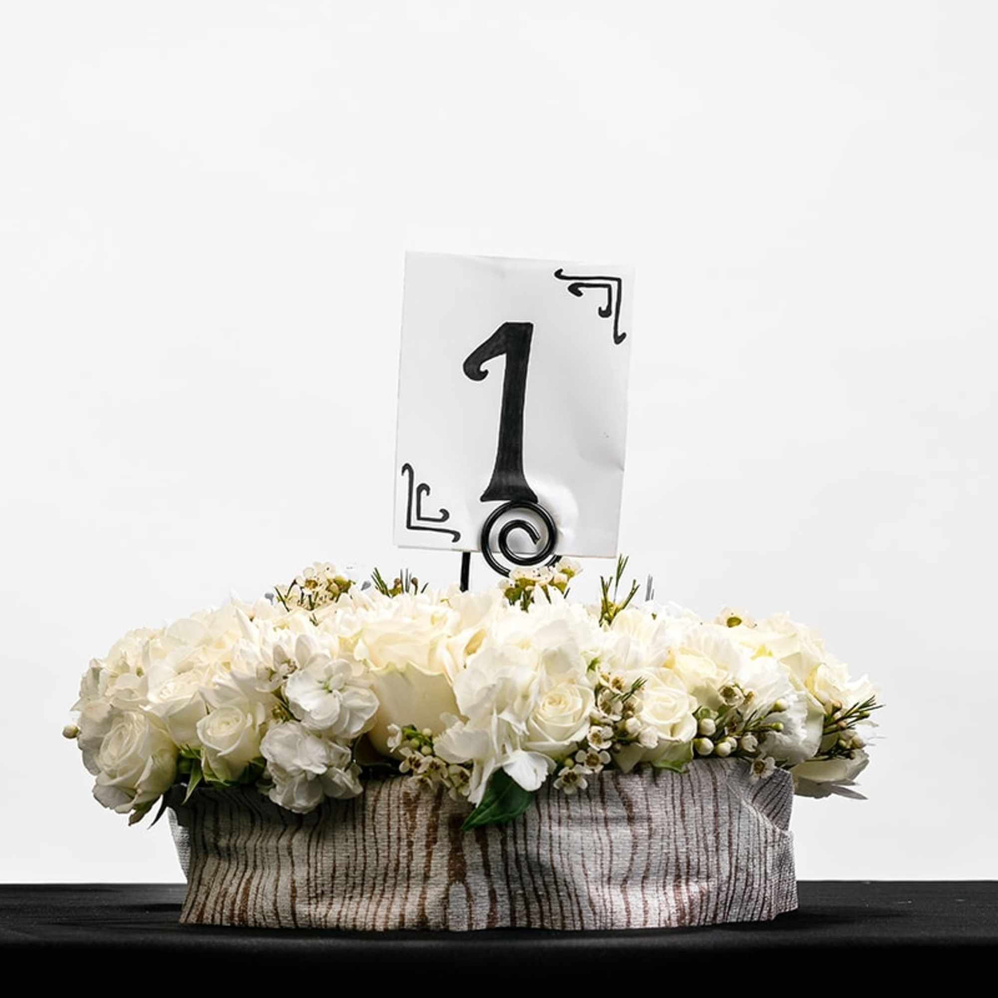 Low white rose centerpiece with table number one card in a fabric-wrapped container