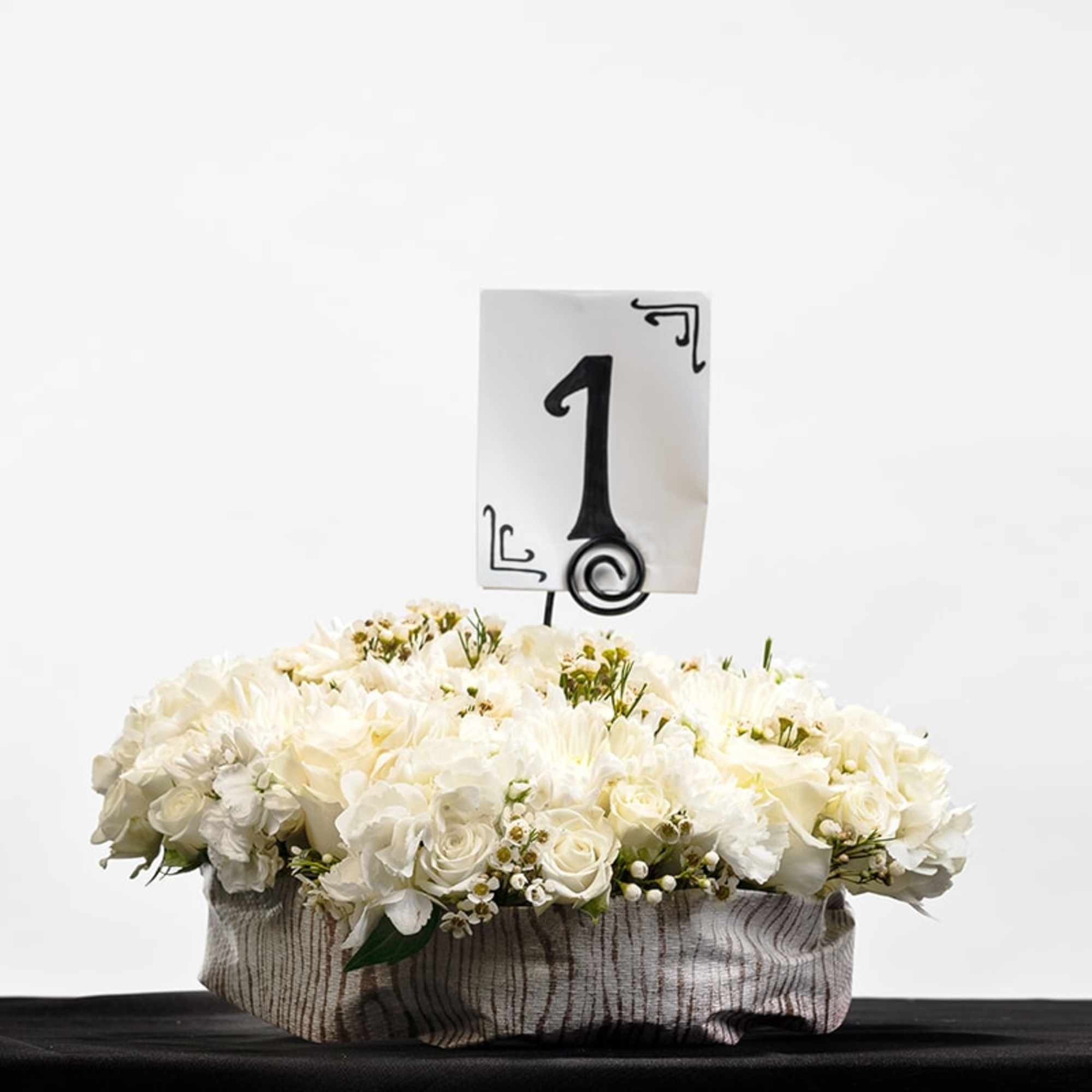 Low round arrangement of white flowers in a fabric-wrapped container with a table number one card