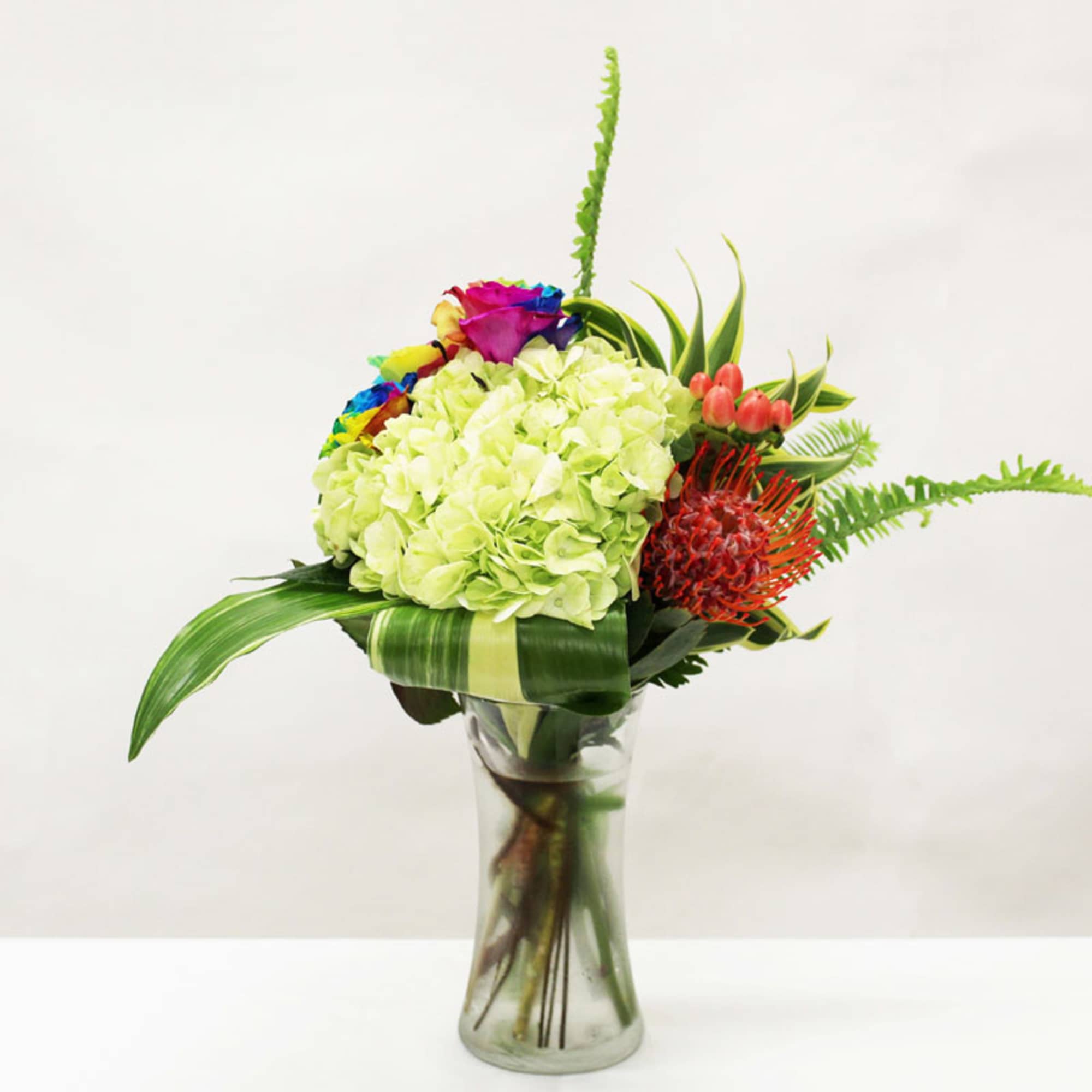 Tall glass vase with green hydrangea, rainbow roses, and orange pincushion protea