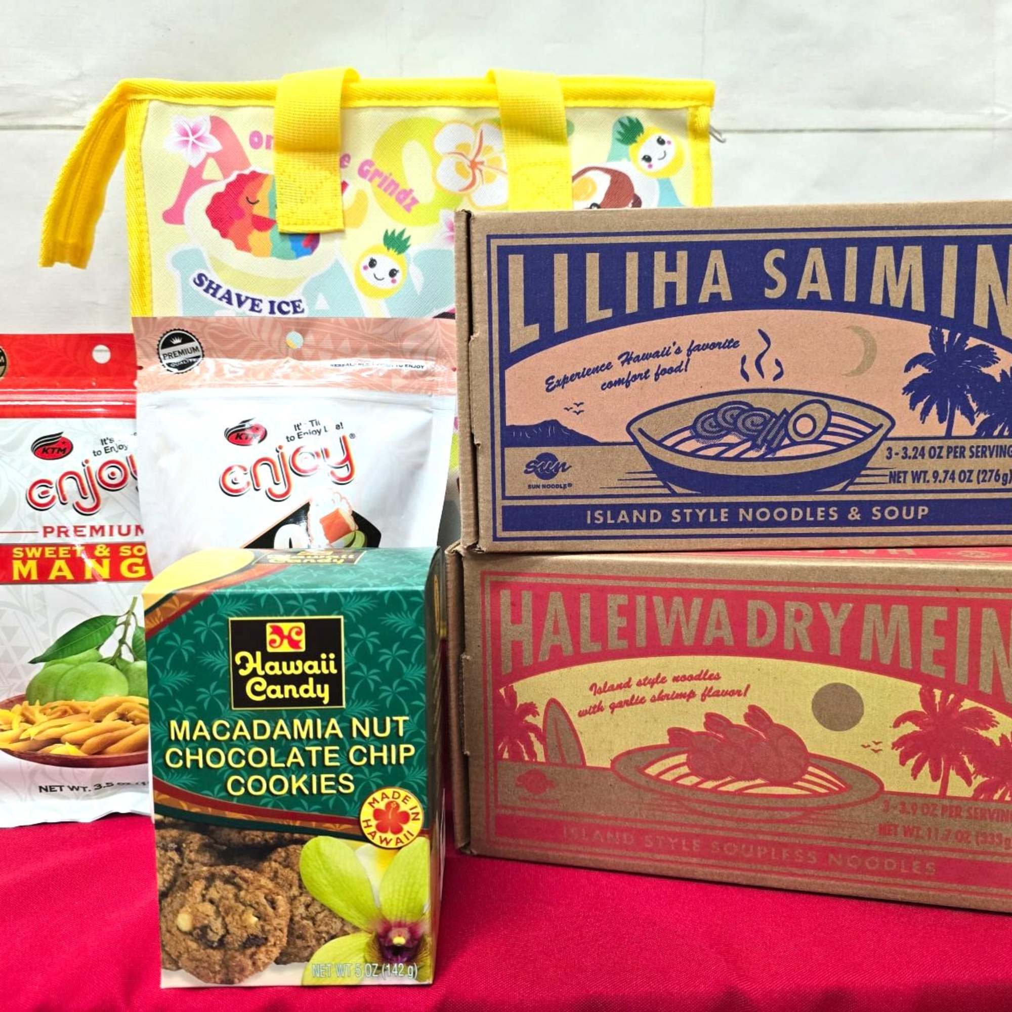 Hawaiian snack gift set with noodles, cookies, dried mango, and yellow insulated tote bag on red cloth
