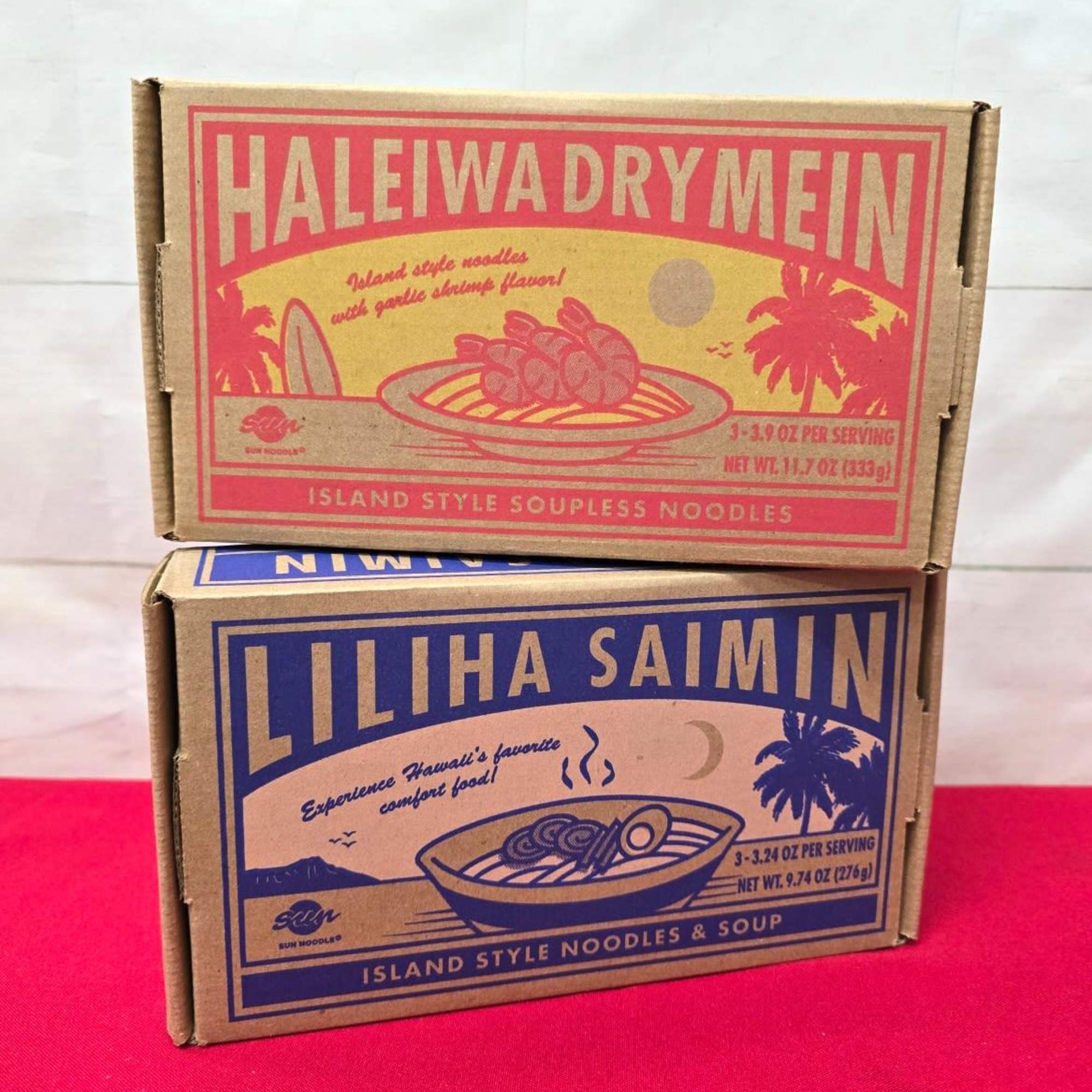 Two stacked cardboard boxes of Hawaiian-style noodle kits on a red cloth surface