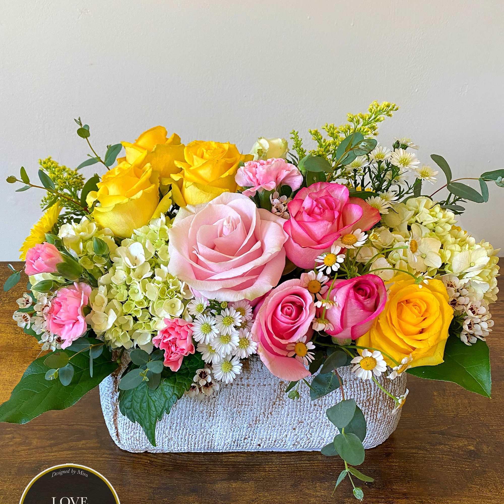 Low arrangement of pink and yellow roses with hydrangeas and daisies in a rectangular white container