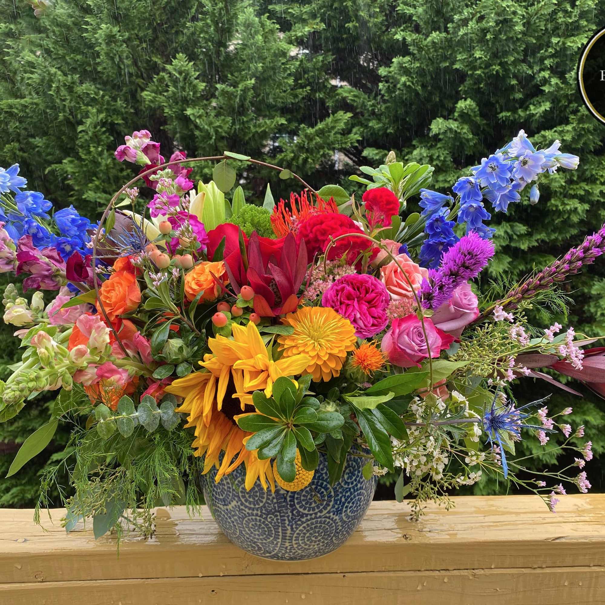 Wide mixed arrangement of bright sunflowers, roses, delphinium and other flowers in a blue pot
