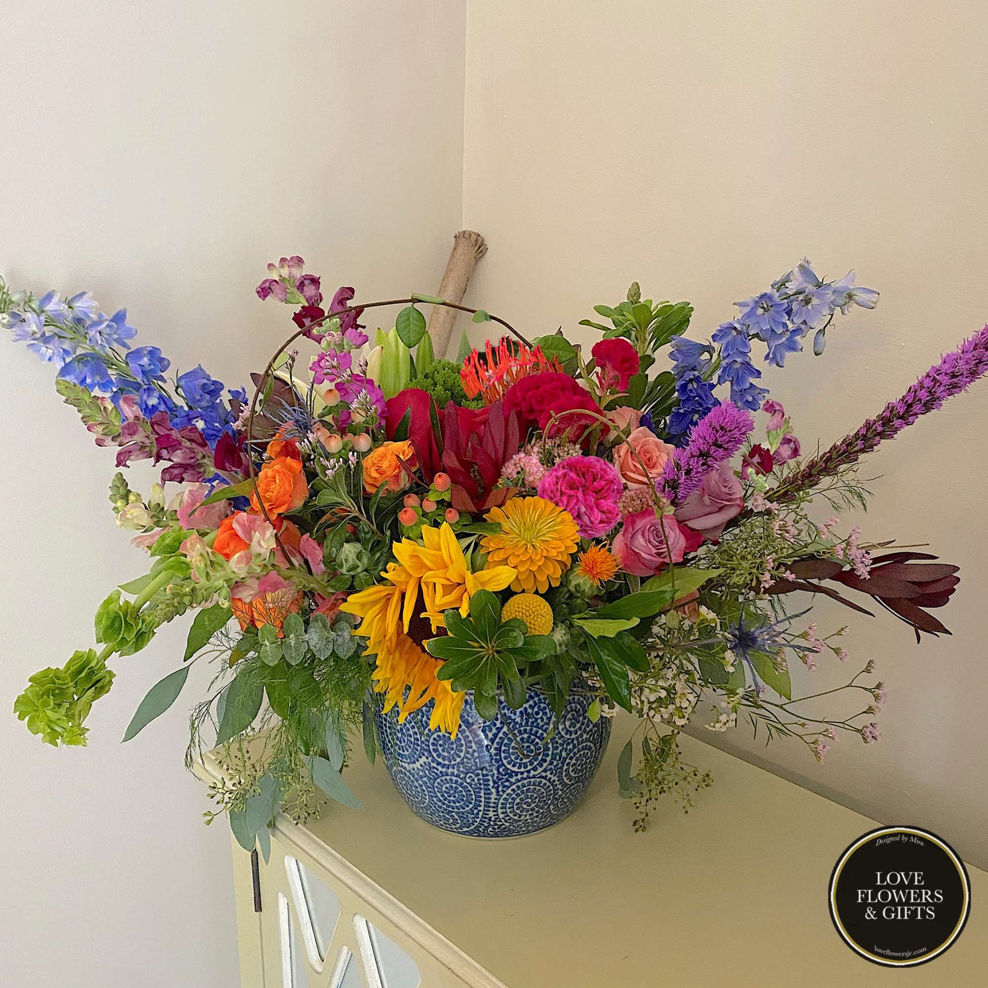 Large mixed-color flower arrangement with sunflowers, roses, and delphinium in a blue patterned ceramic pot