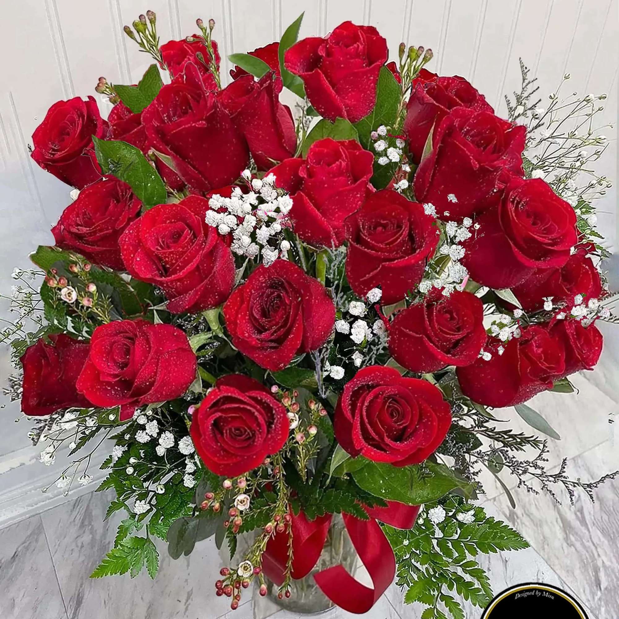 Tall bouquet of red roses with white filler flowers in a clear glass vase with a red ribbon