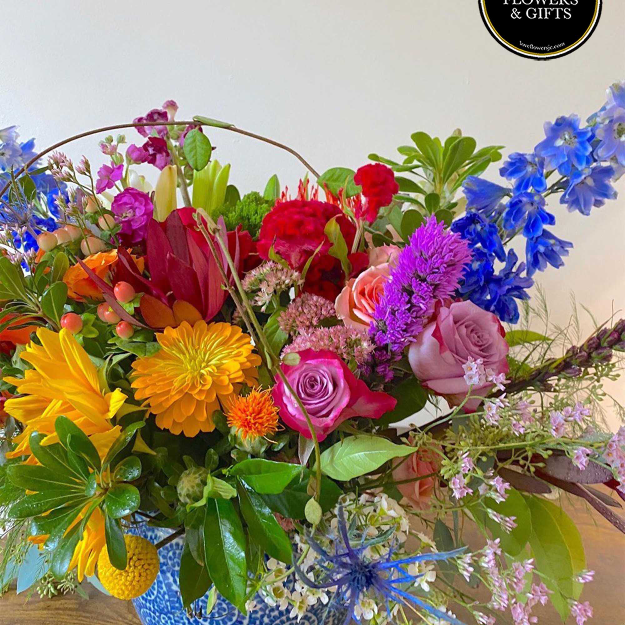 Colorful mixed flower arrangement with roses, delphinium, and daisies in a blue patterned ceramic pot.