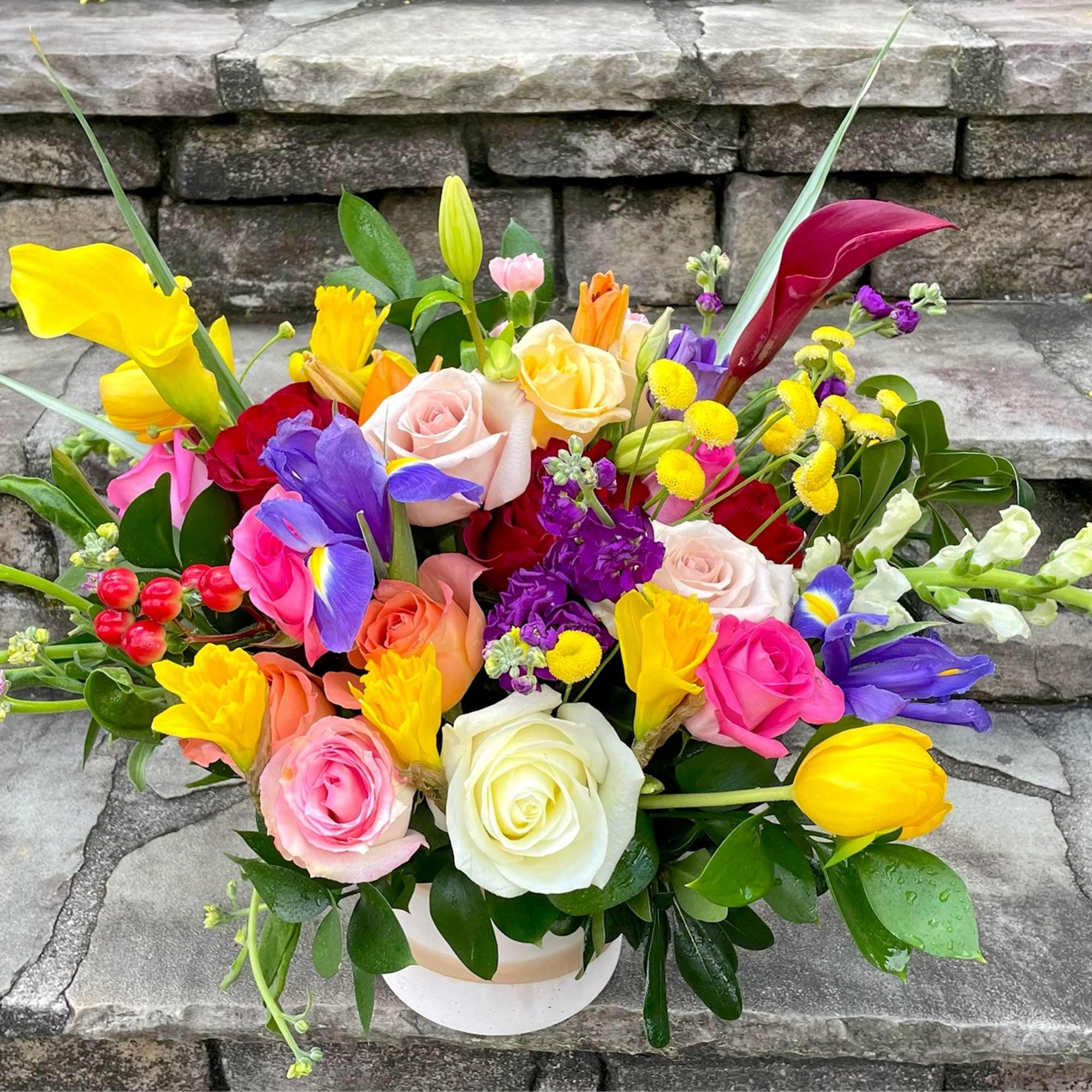 Low round arrangement of colorful roses, tulips, irises, and other blooms in a white container.