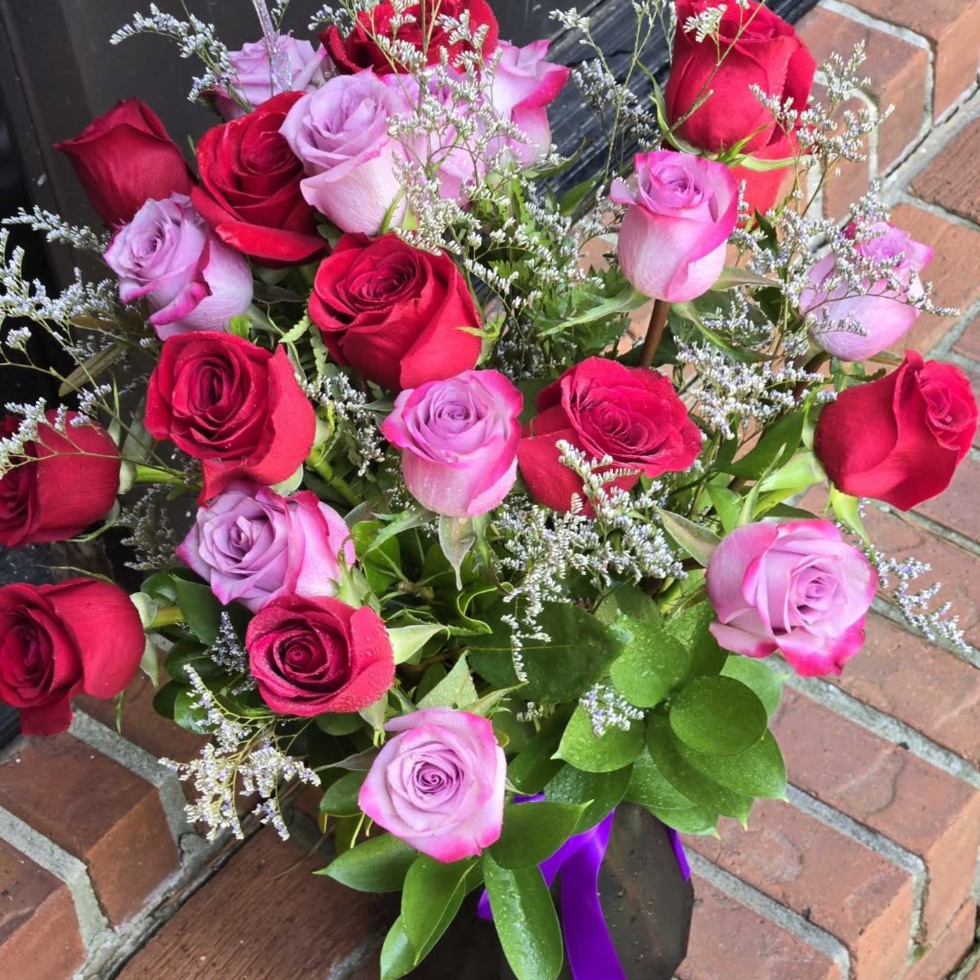 Arrangement of red and pink roses with white filler flowers in a black vase with a purple ribbon
