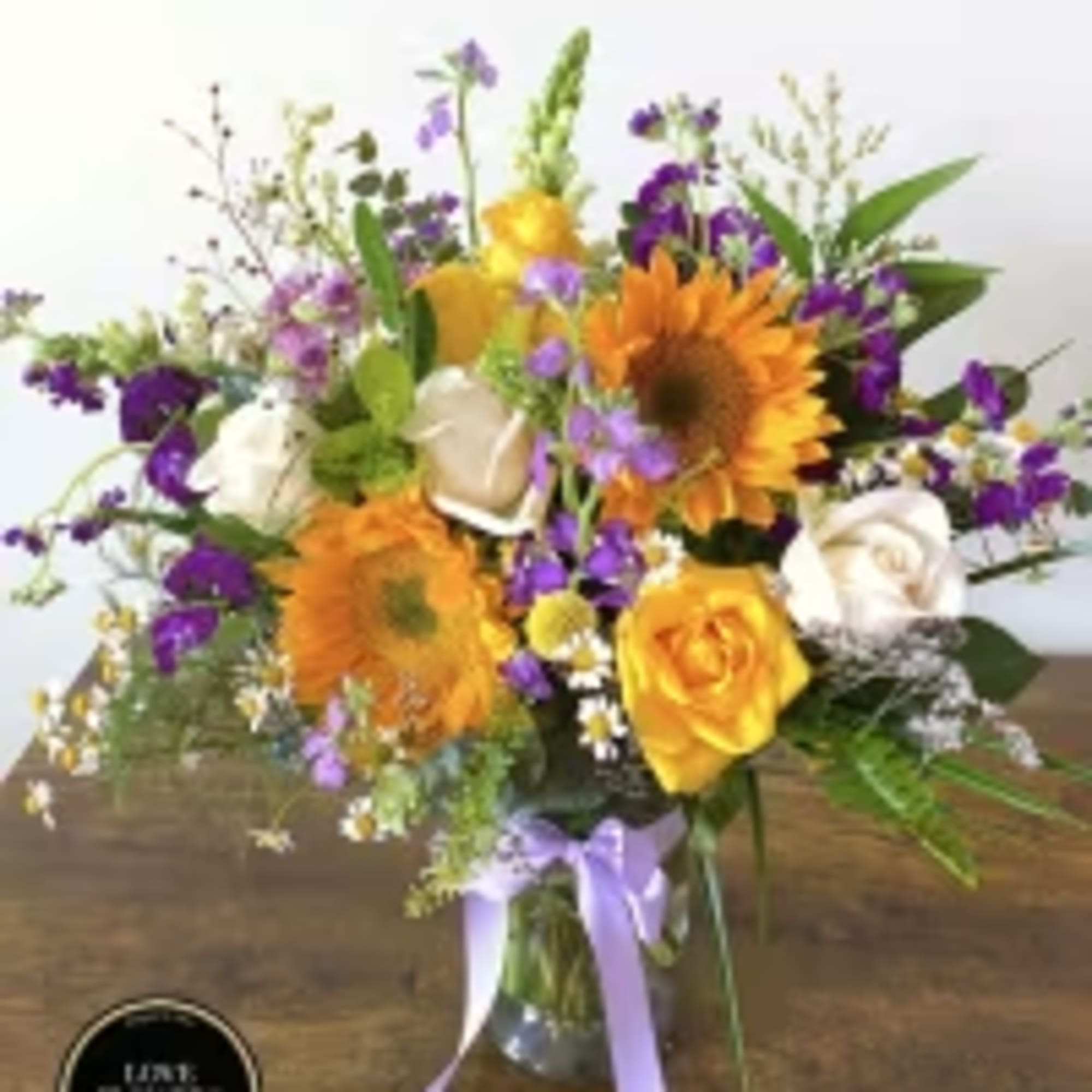Mixed arrangement of yellow sunflowers, roses, and purple flowers in a clear vase with a lavender bow