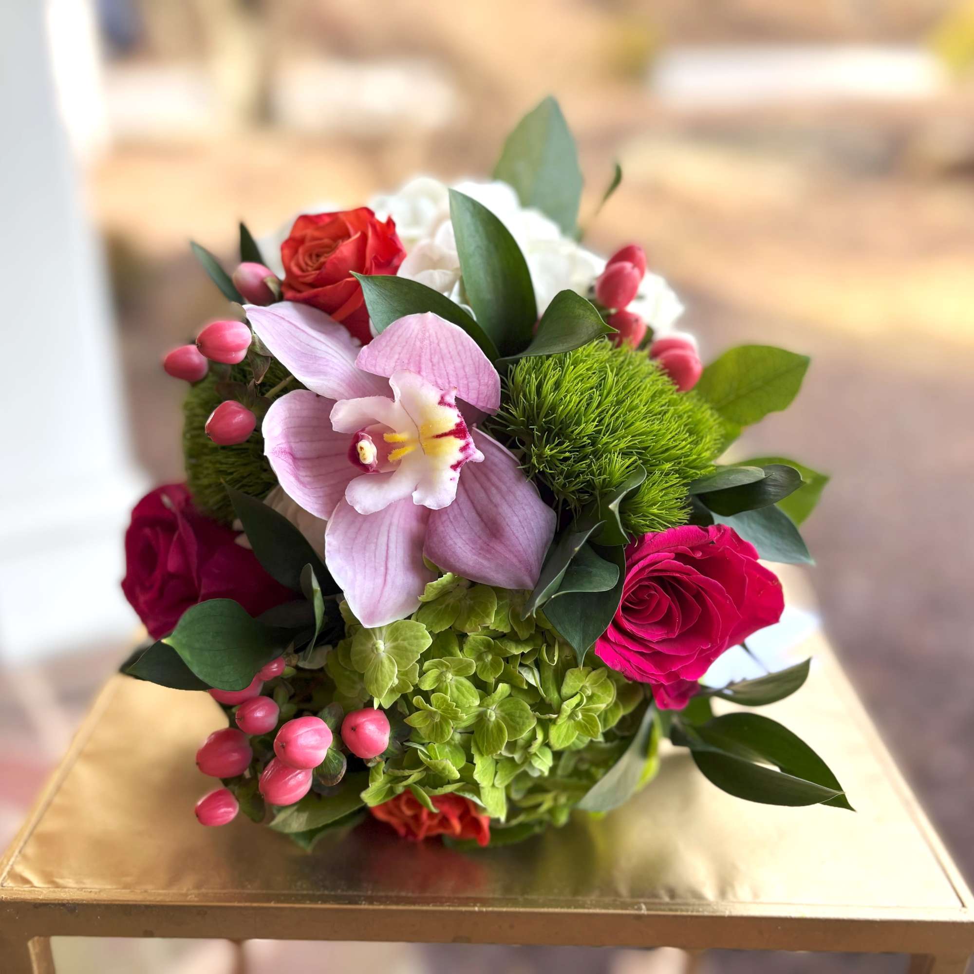 Low arrangement with pink orchid, bright roses, and green hydrangea set on a gold surface
