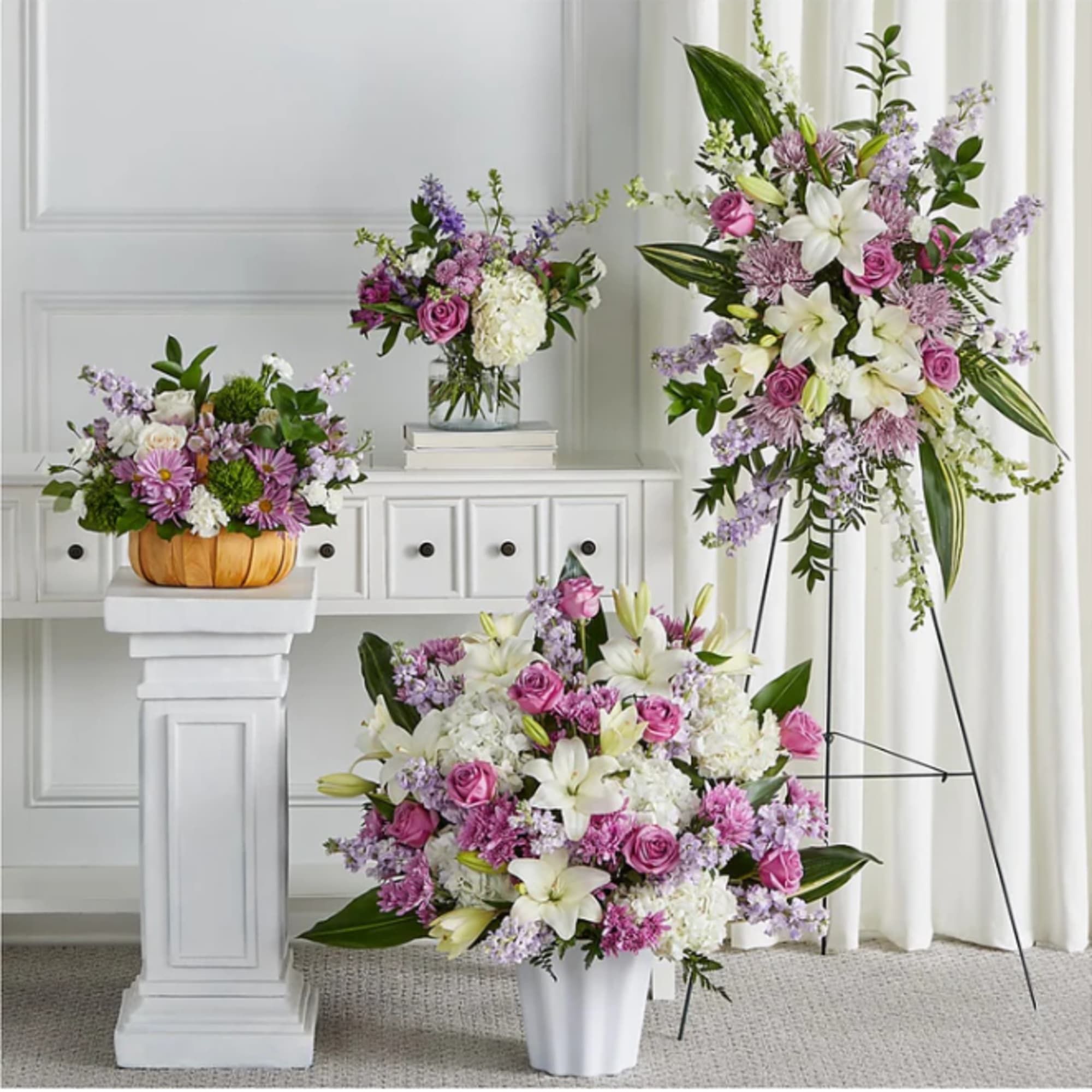 Four floral arrangements in pink, white, and lavender tones displayed on pedestals and stands.