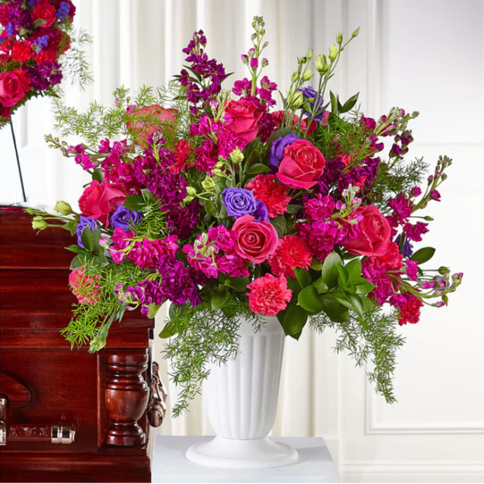 Bright pink and purple floral arrangement in a white vase beside a casket