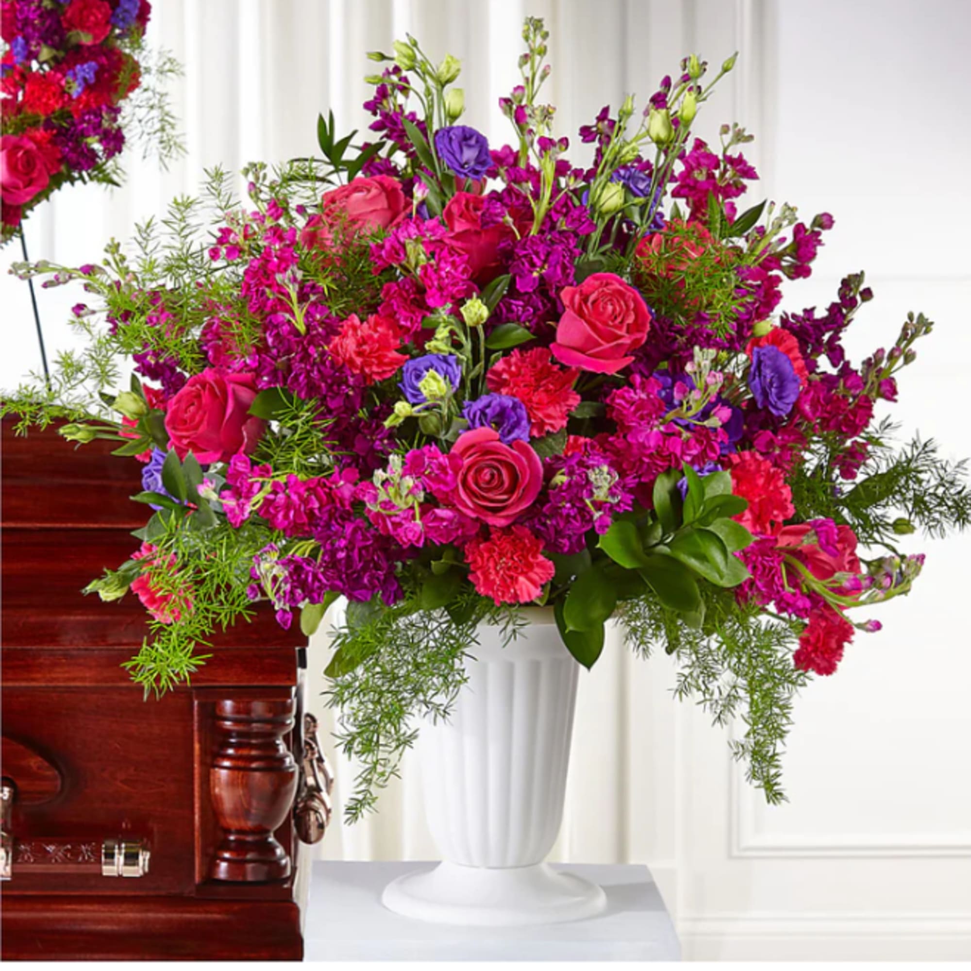 Bright pink and purple floral arrangement in a white urn beside a casket