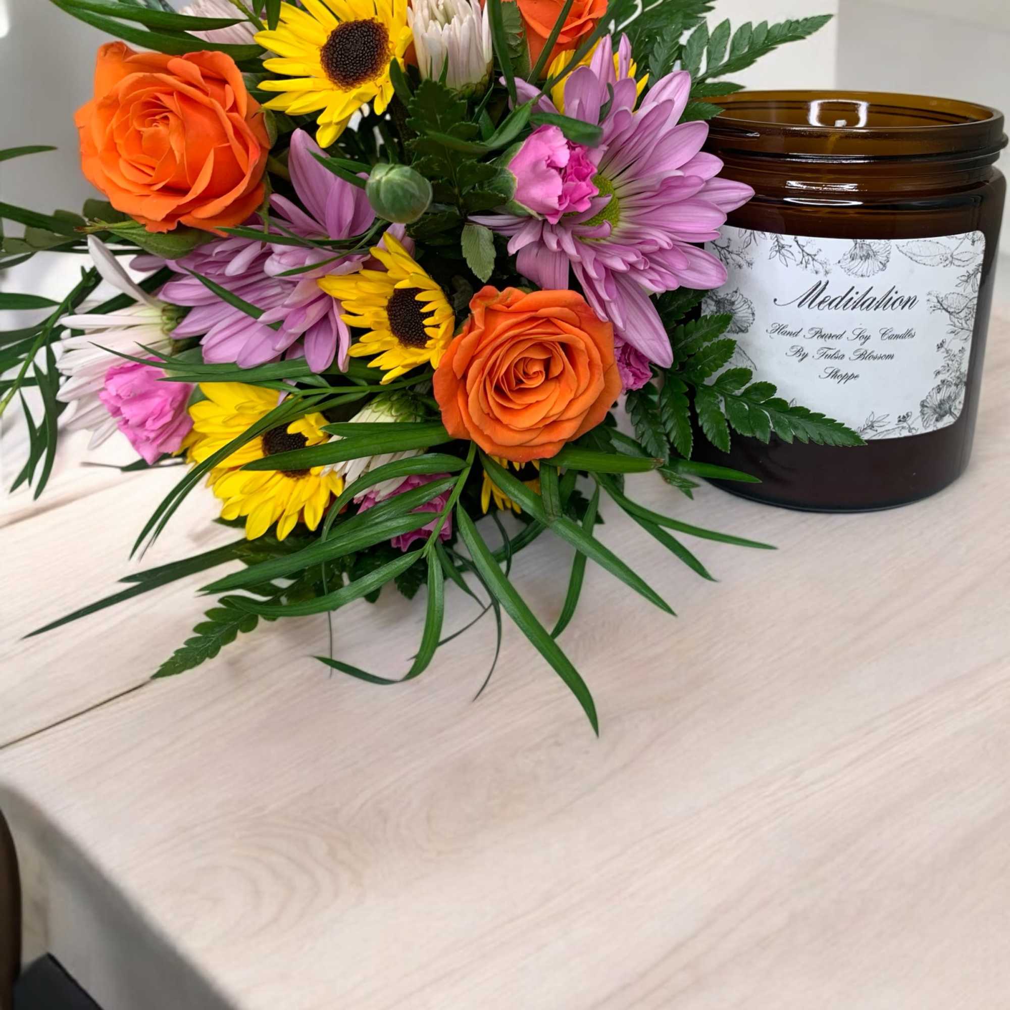 Bouquet of orange roses, sunflowers, and purple daisies beside a brown candle jar