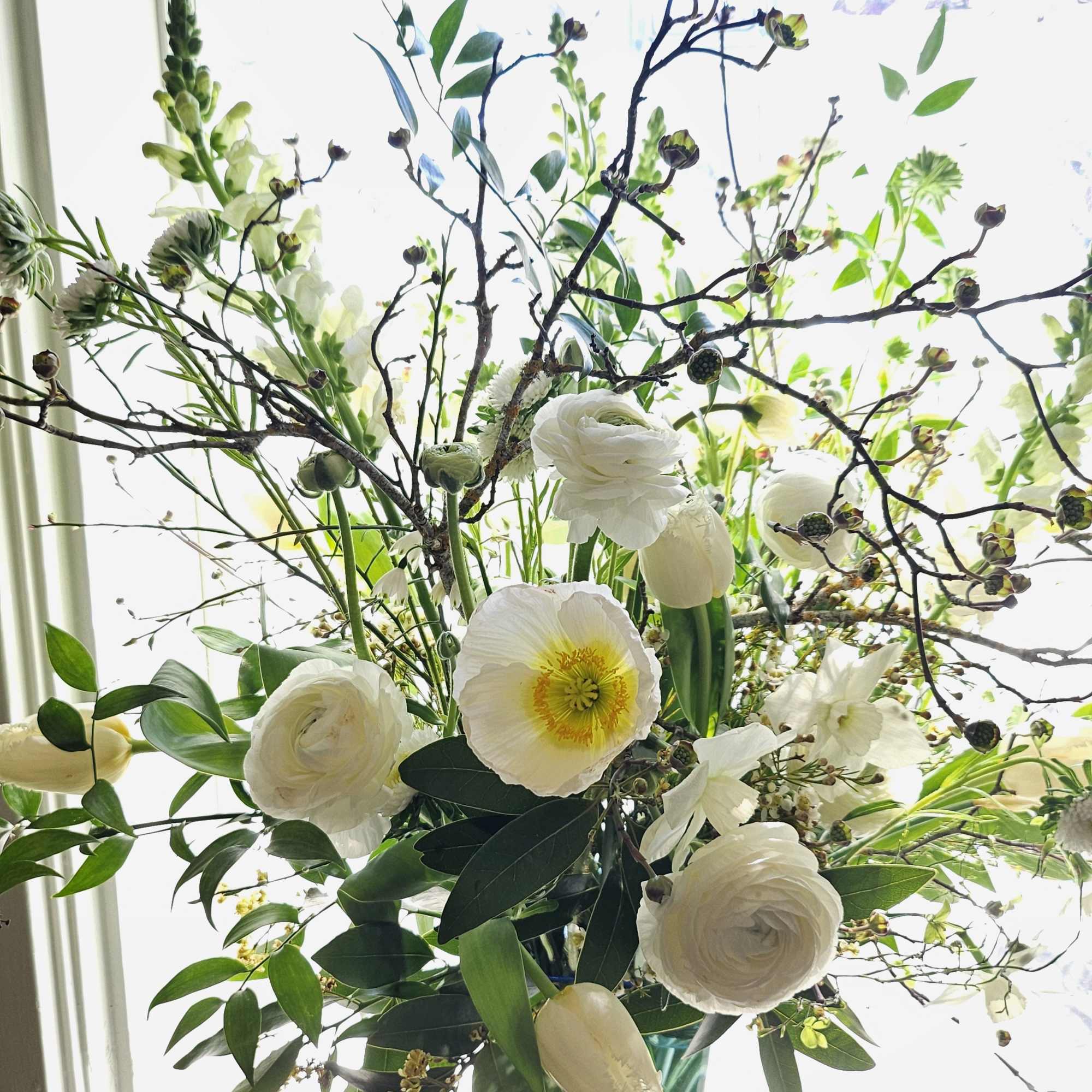Tall arrangement of white flowers and twiggy branches in a blue glass vase by a bright window