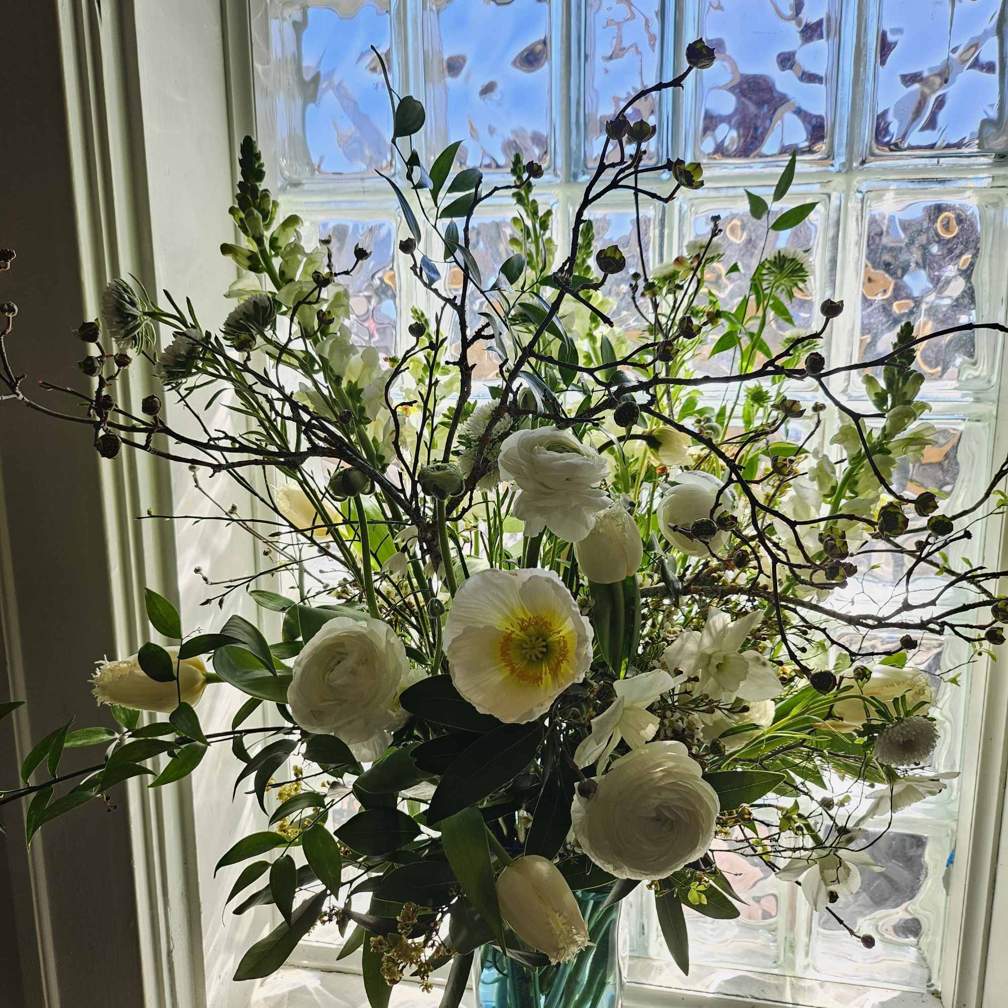 Tall arrangement of white flowers with branches in a blue glass vase on a windowsill