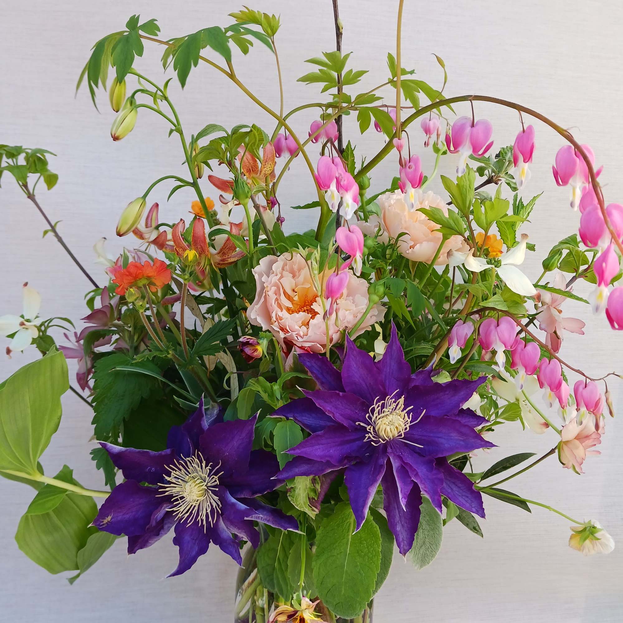 Tall mixed spring bouquet with purple, pink, peach, and white flowers in a clear glass vase